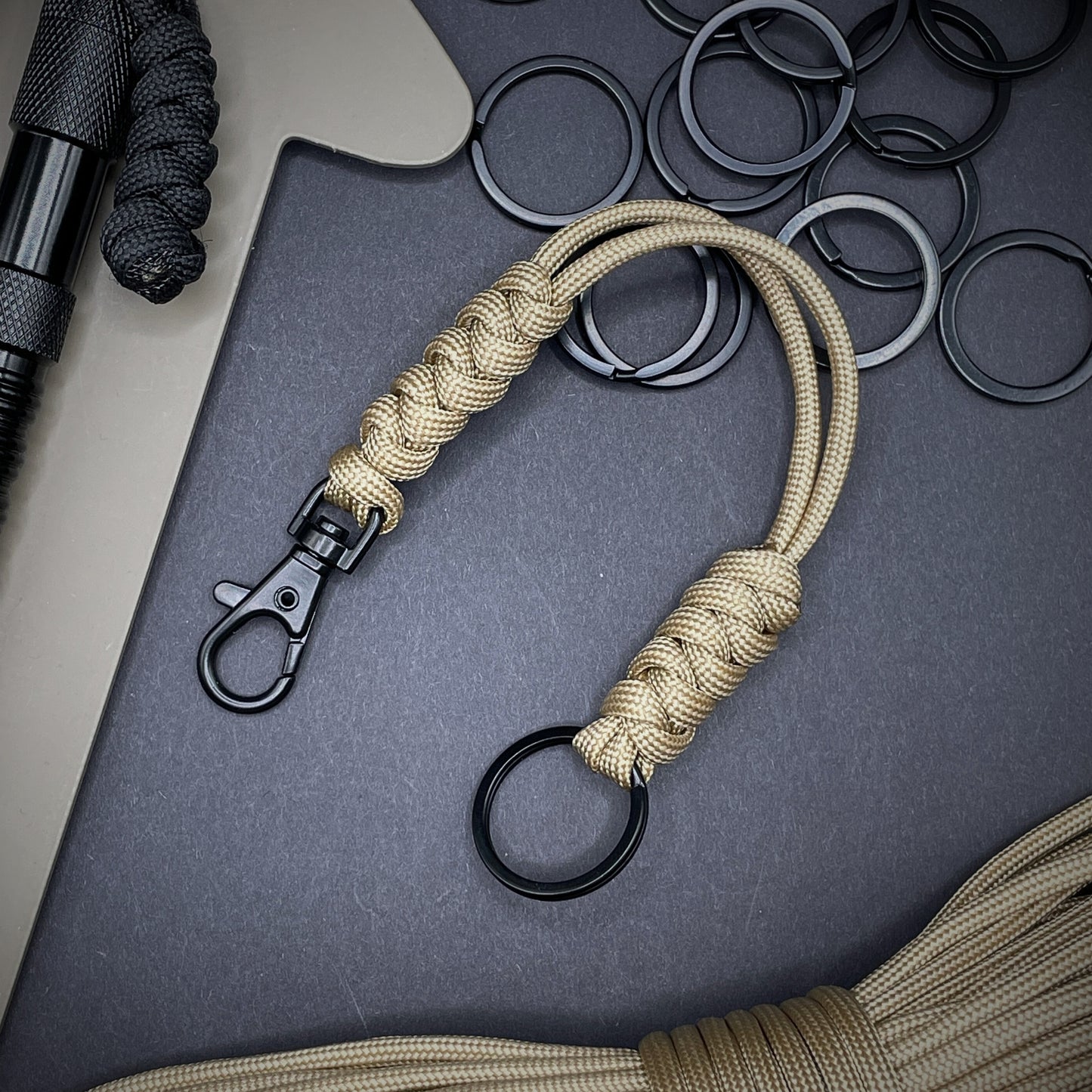 Khaki 550 Paracord snake knot lanyard with black split ring and lobster clasp, displayed on a dark textured background with EDC gear in the background.