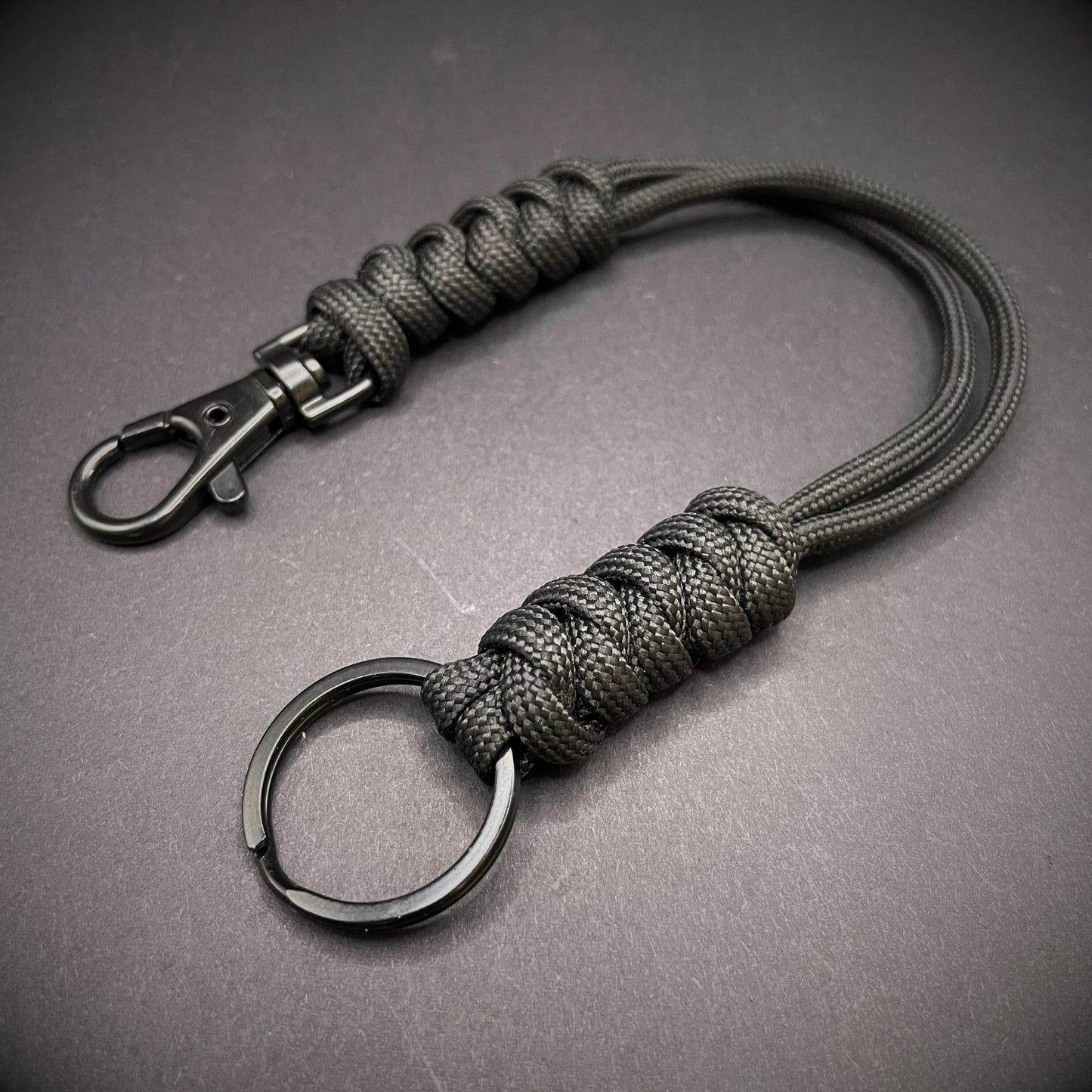 Black 550 Paracord snake knot lanyard with black split ring and lobster clasp, displayed on a dark textured background.