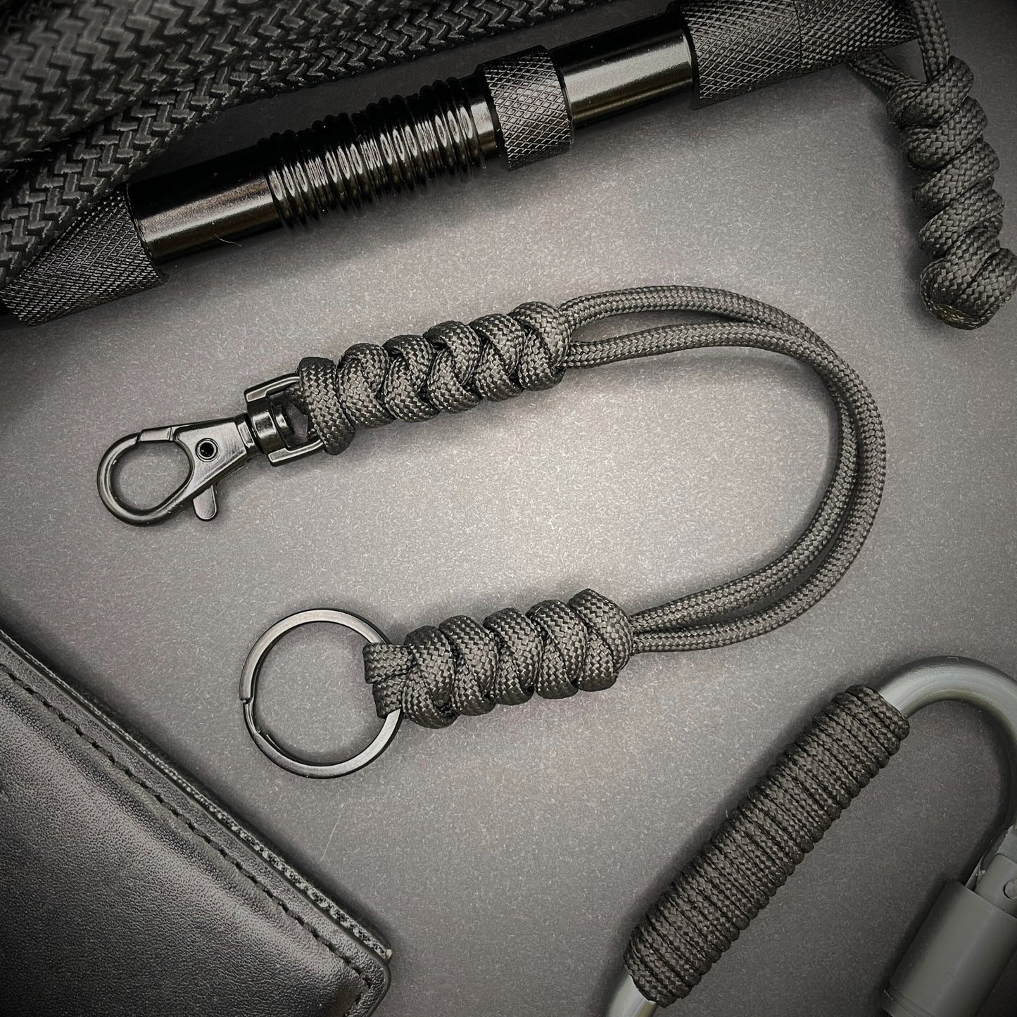 Black 550 Paracord snake knot lanyard with black split ring and lobster clasp, displayed on a dark textured background with EDC gear in the background.