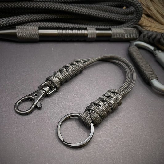 Black 550 Paracord snake knot lanyard with black split ring and lobster clasp, displayed on a dark textured background with EDC gear in the background.