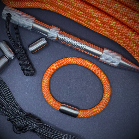 Bright orange braided rope bracelet with a stainless steel cylindrical clasps and black whipping detail, styled on a dark background with EDC gear in the backdrop.