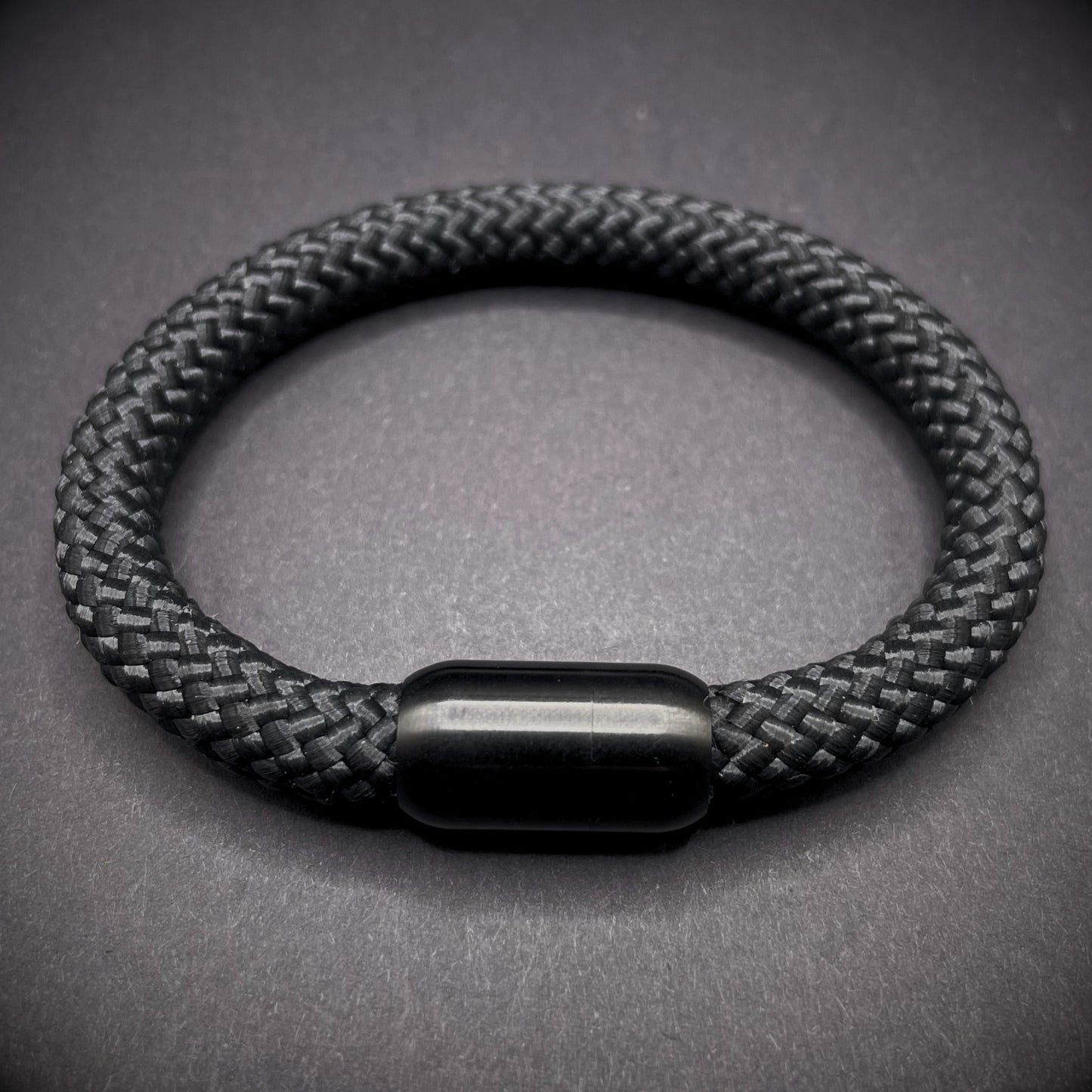 Black braided rope bracelet with a sleek cylindrical black metal clasp, displayed on a dark textured background.