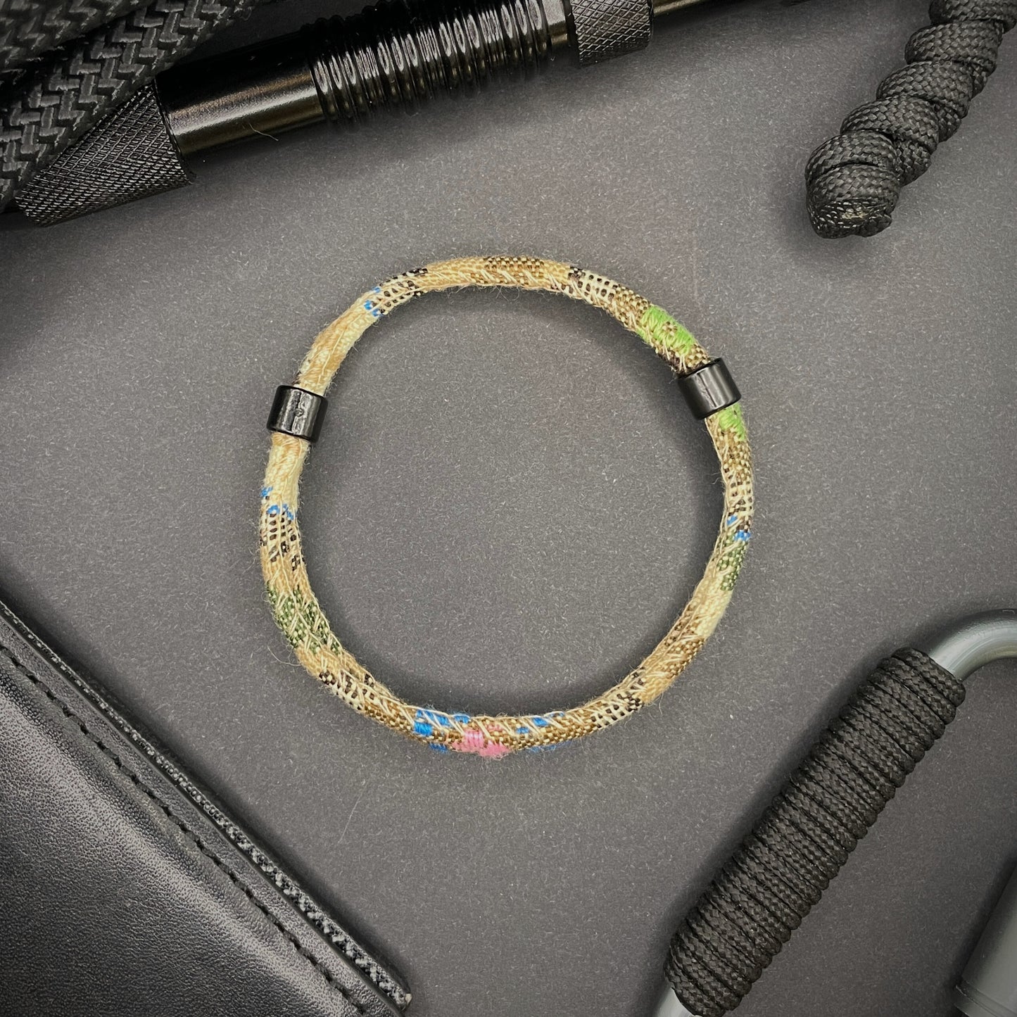 Khaki Boho rope bracelet with black sliding clasps, displayed on a dark grey surface with EDC gear in the background.