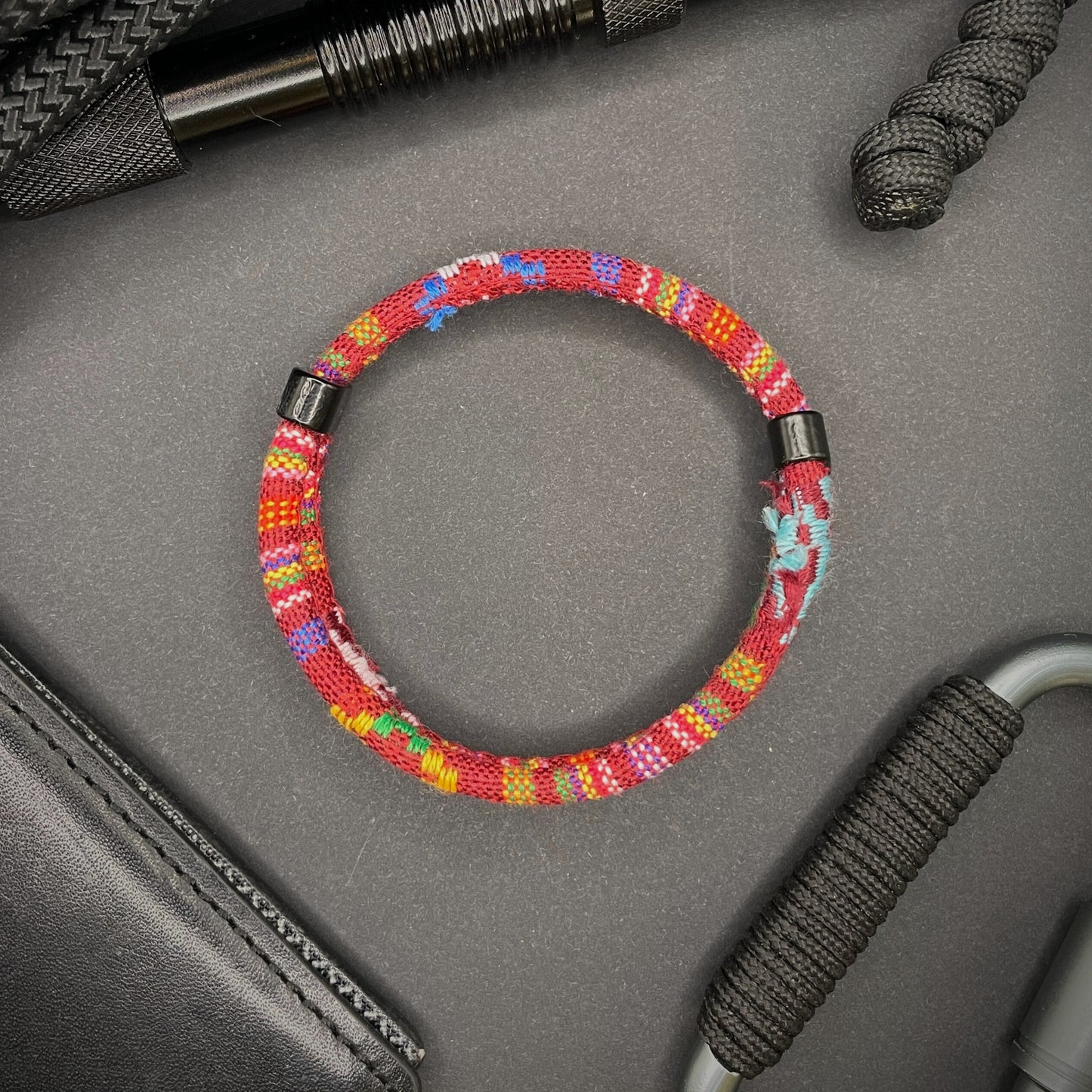 Red Boho rope bracelet with black sliding clasps, displayed on a dark grey surface with EDC gear in the background.