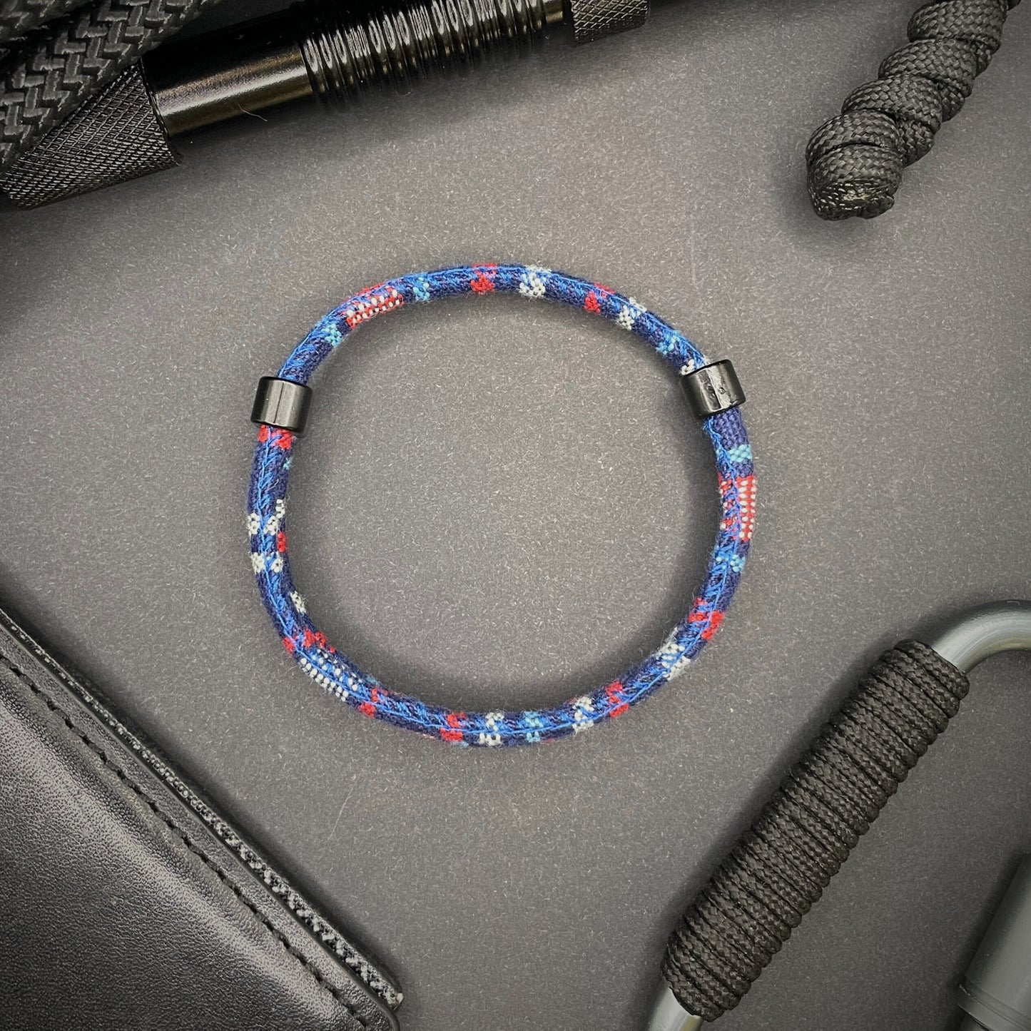 Dark Blue and red Boho rope bracelet with black sliding clasps, displayed on a dark grey surface with EDC gear in the background.