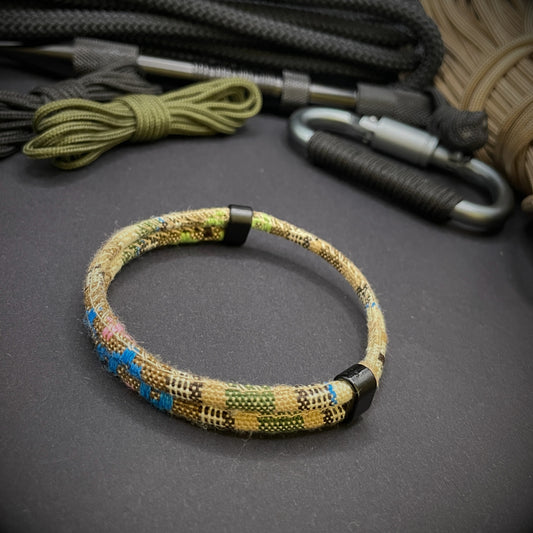 Khaki Boho rope bracelet with black sliding clasps, displayed on a dark grey surface with EDC gear in the background.