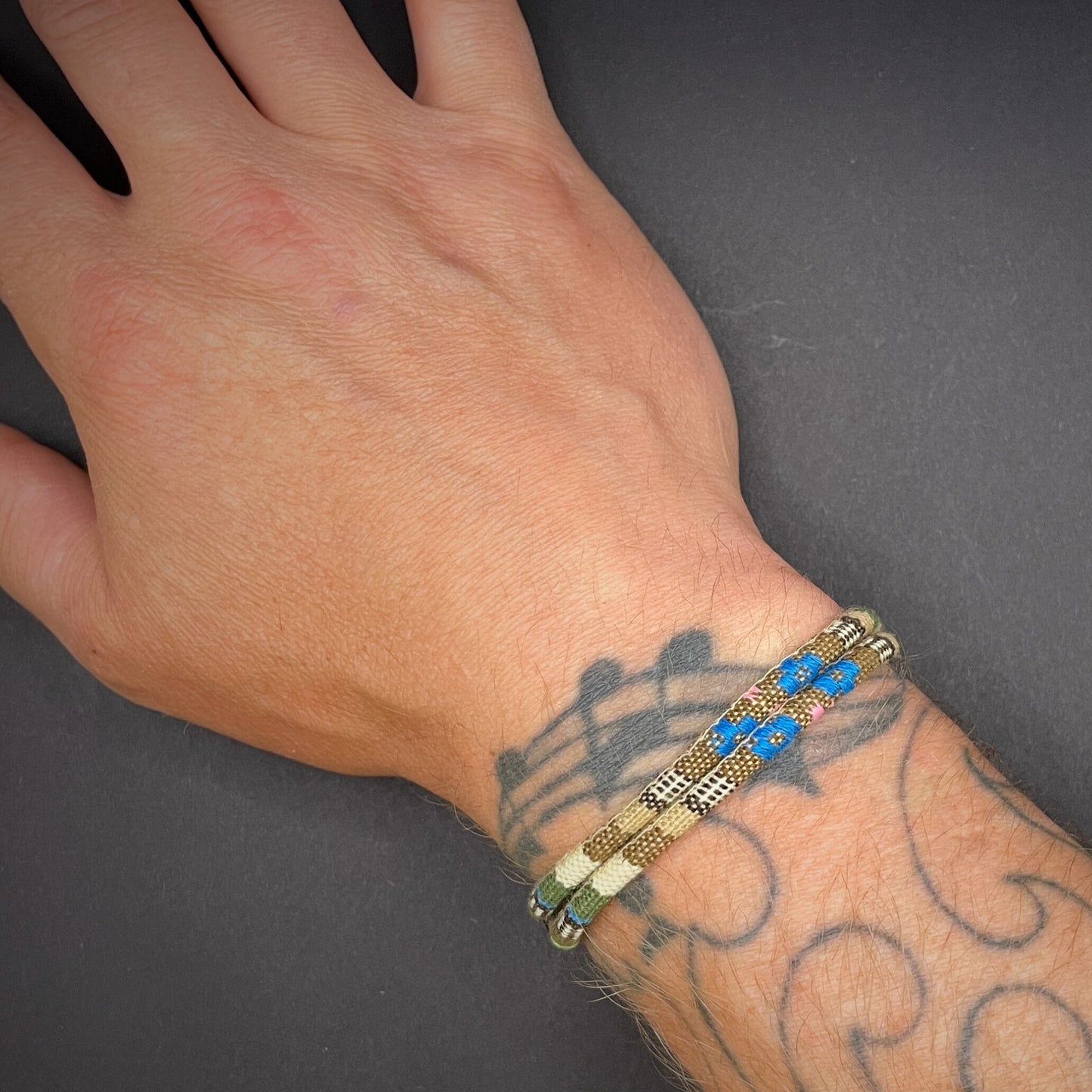 Khaki Boho rope bracelet with black sliding clasps, displayed on a tattooed wrist over a dark grey surface.