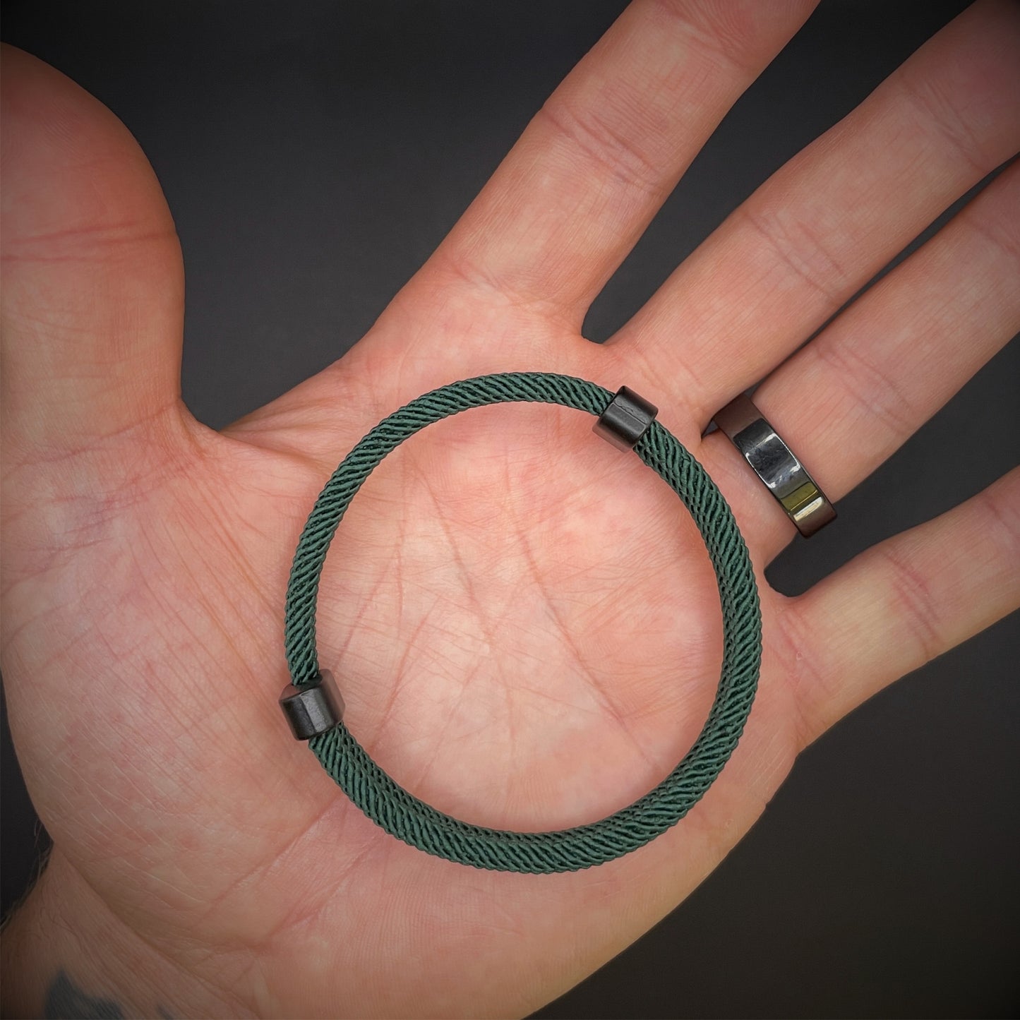 Forrest Green Milan rope bracelet with black sliding clasps, displayed in an open palmed hand over a dark grey surface.