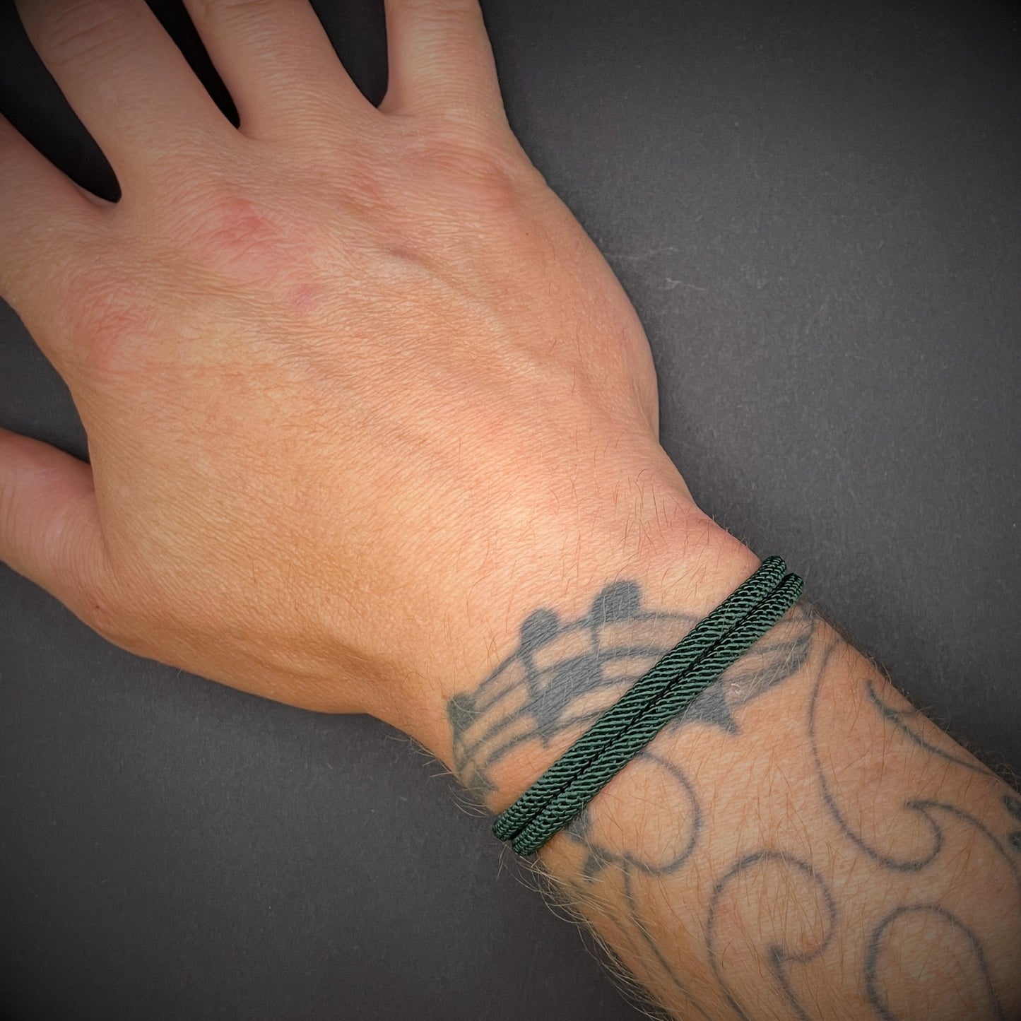 Forrest Green Milan rope bracelet with black sliding clasps, displayed on a tattooed wrist over a dark grey surface.