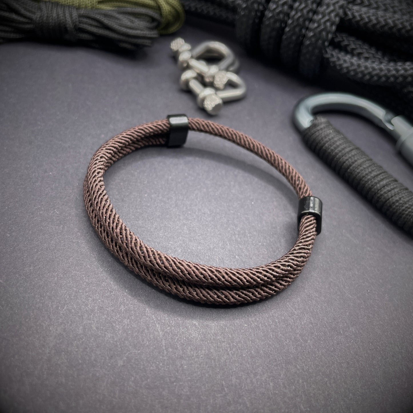 Coffee Milan rope bracelet with black sliding clasps, displayed on a dark grey surface with EDC gear in the background.