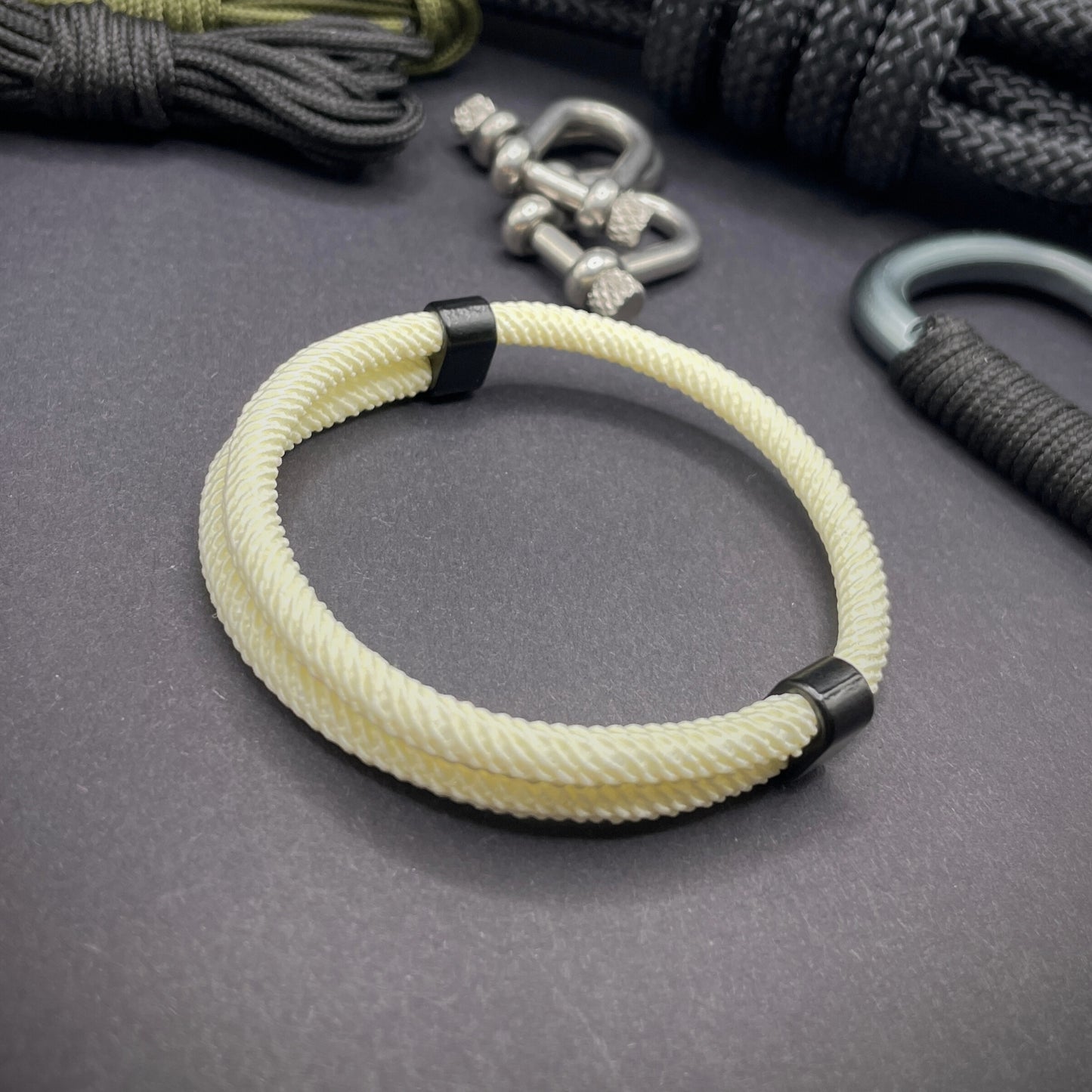 Cream Milan rope bracelet with black sliding clasps, displayed on a dark grey surface with EDC gear in the background.