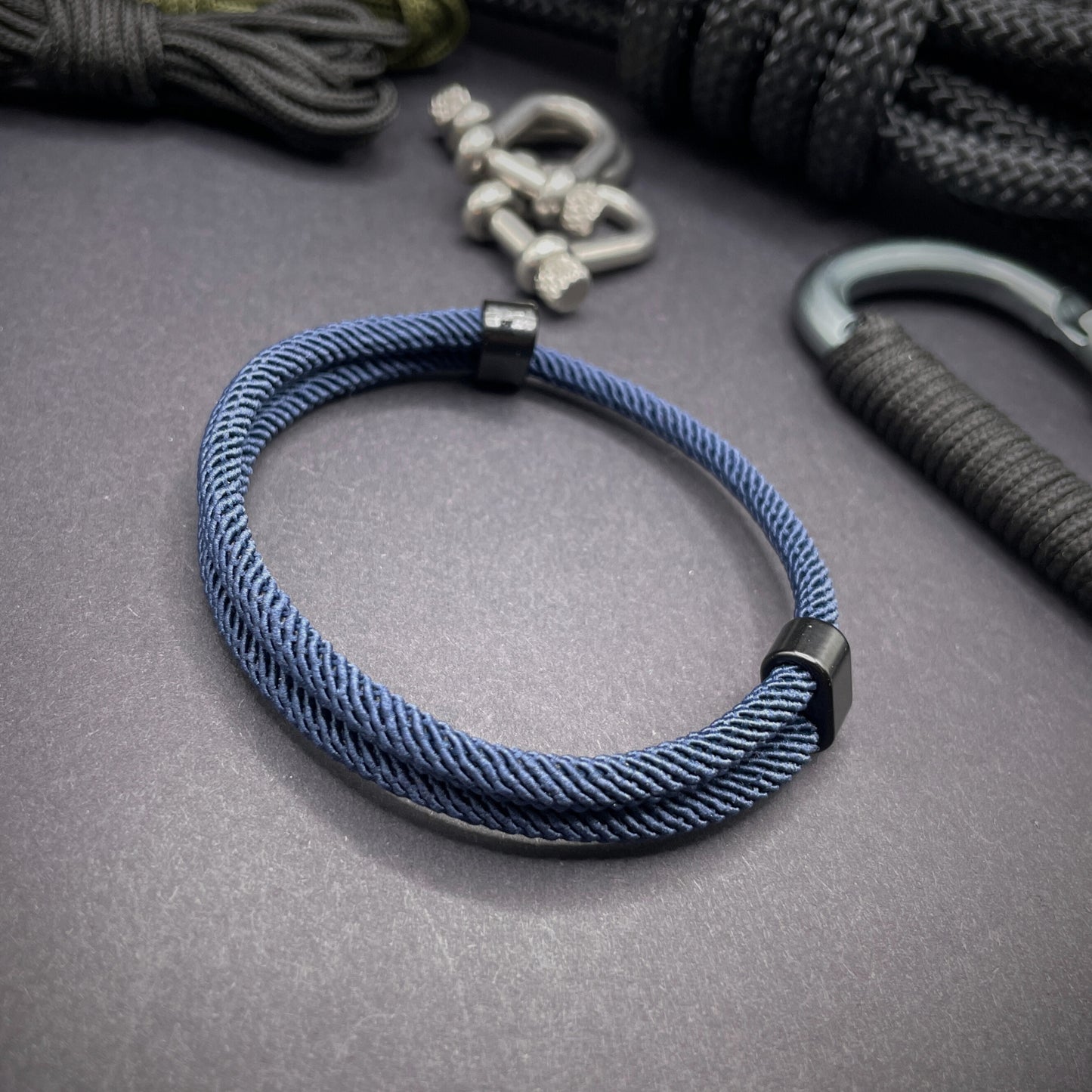 Dark Blue Milan rope bracelet with black sliding clasps, displayed on a dark grey surface with EDC gear in the background.