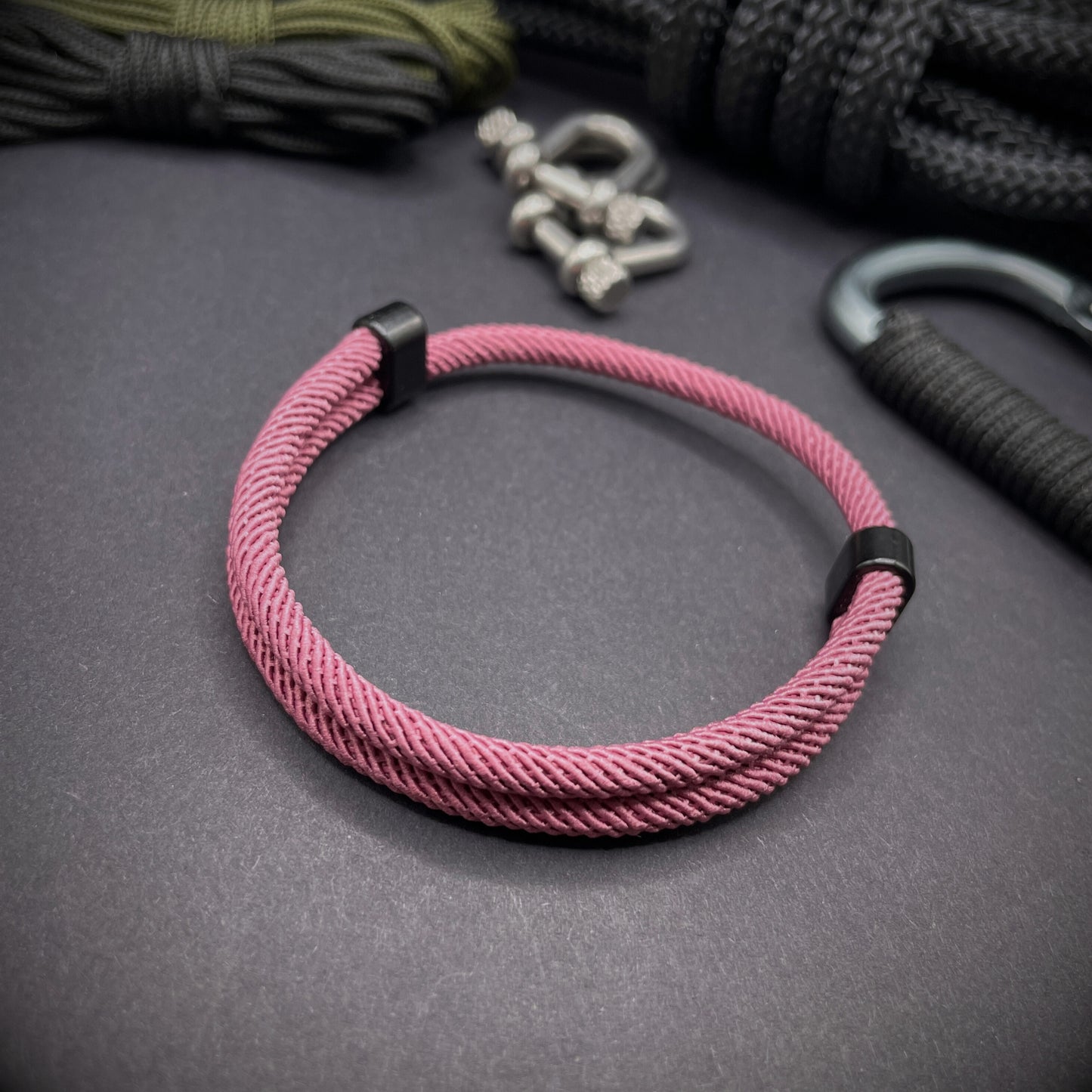 Pink Milan rope bracelet with black sliding clasps, displayed on a dark grey surface with EDC gear in the background.