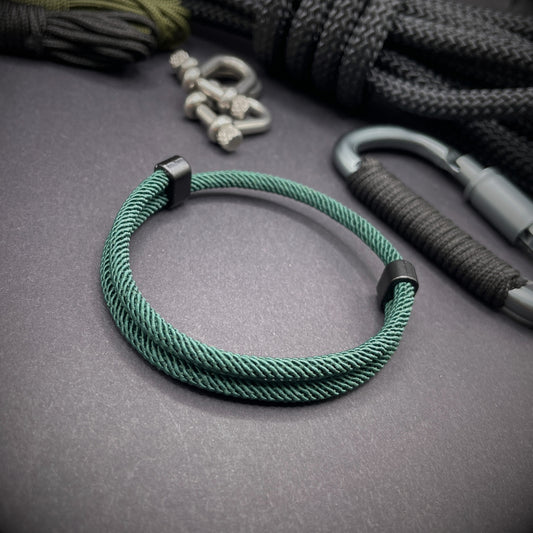 Forrest Green Milan rope bracelet with black sliding clasps, displayed on a dark grey surface with EDC gear in the background.