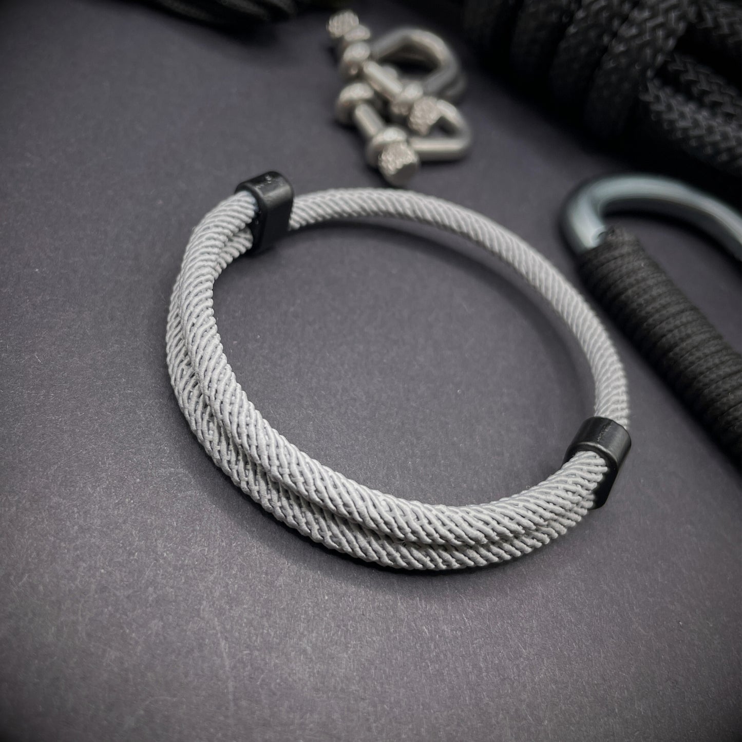 Grey Milan rope bracelet with black sliding clasps, displayed on a dark grey surface with EDC gear in the background.