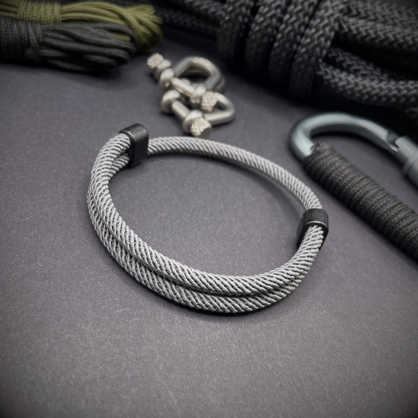 Grey Milan rope bracelet with black sliding clasps, displayed on a dark grey surface with EDC gear in the background.
