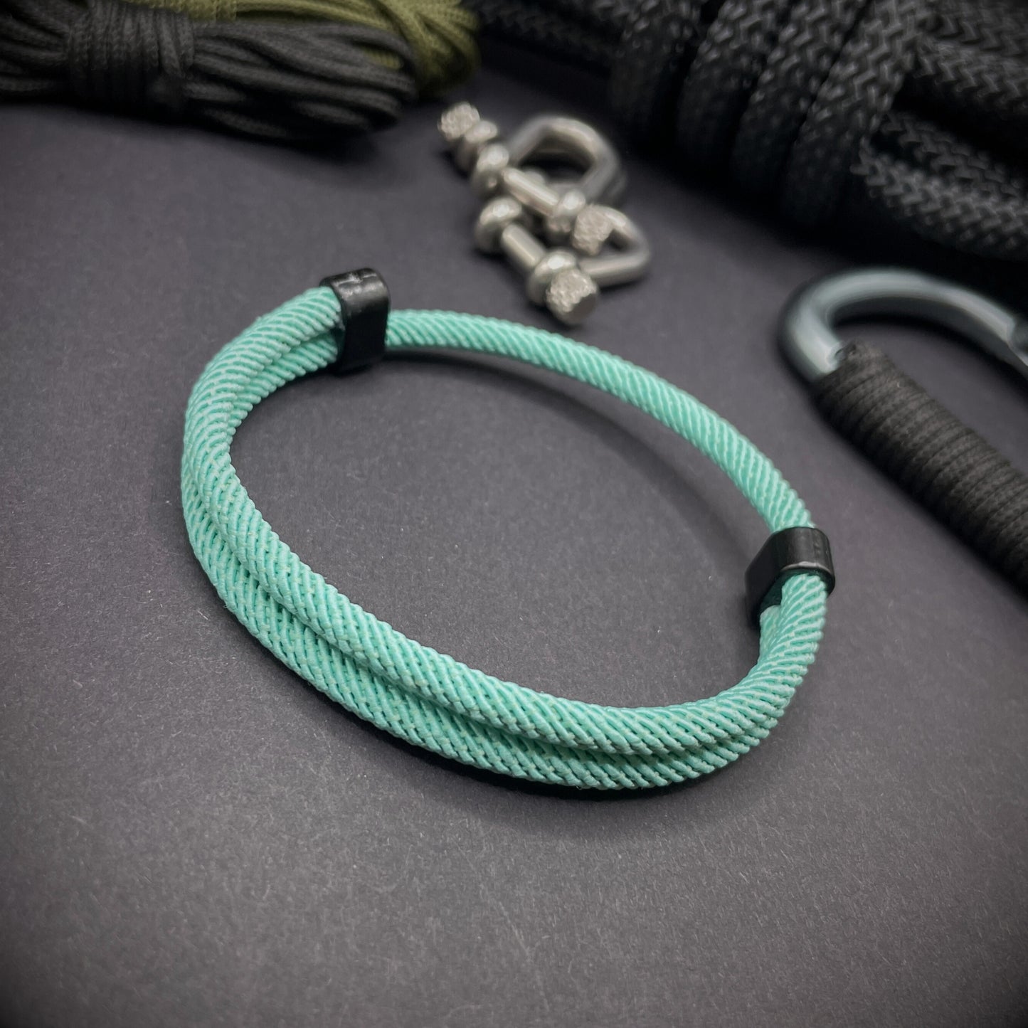 Mint Green Milan rope bracelet with black sliding clasps, displayed on a dark grey surface with EDC gear in the background.