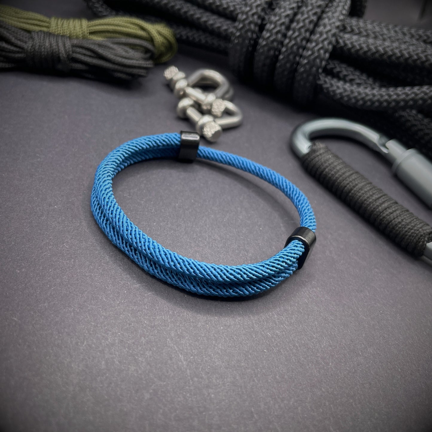 Ocean Blue Milan rope bracelet with black sliding clasps, displayed on a dark grey surface with EDC gear in the background.