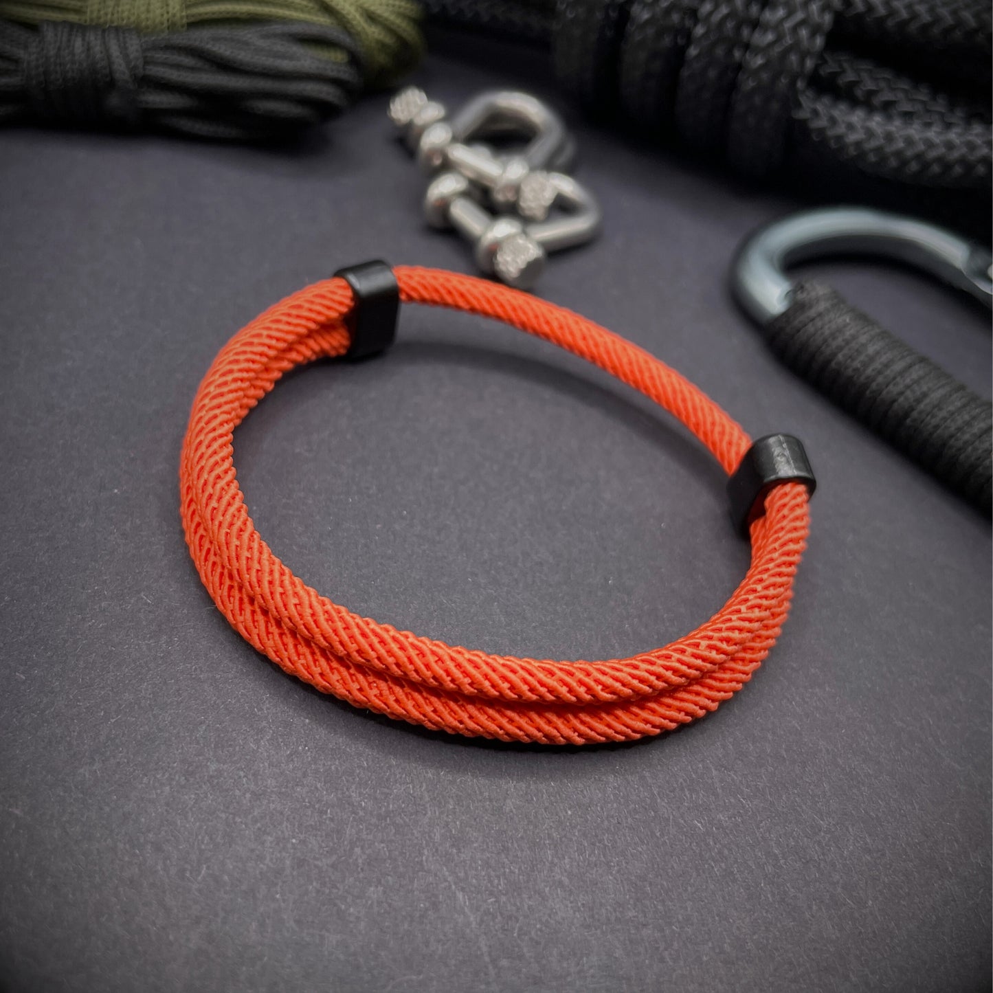 Orange Milan rope bracelet with black sliding clasps, displayed on a dark grey surface with EDC gear in the background.