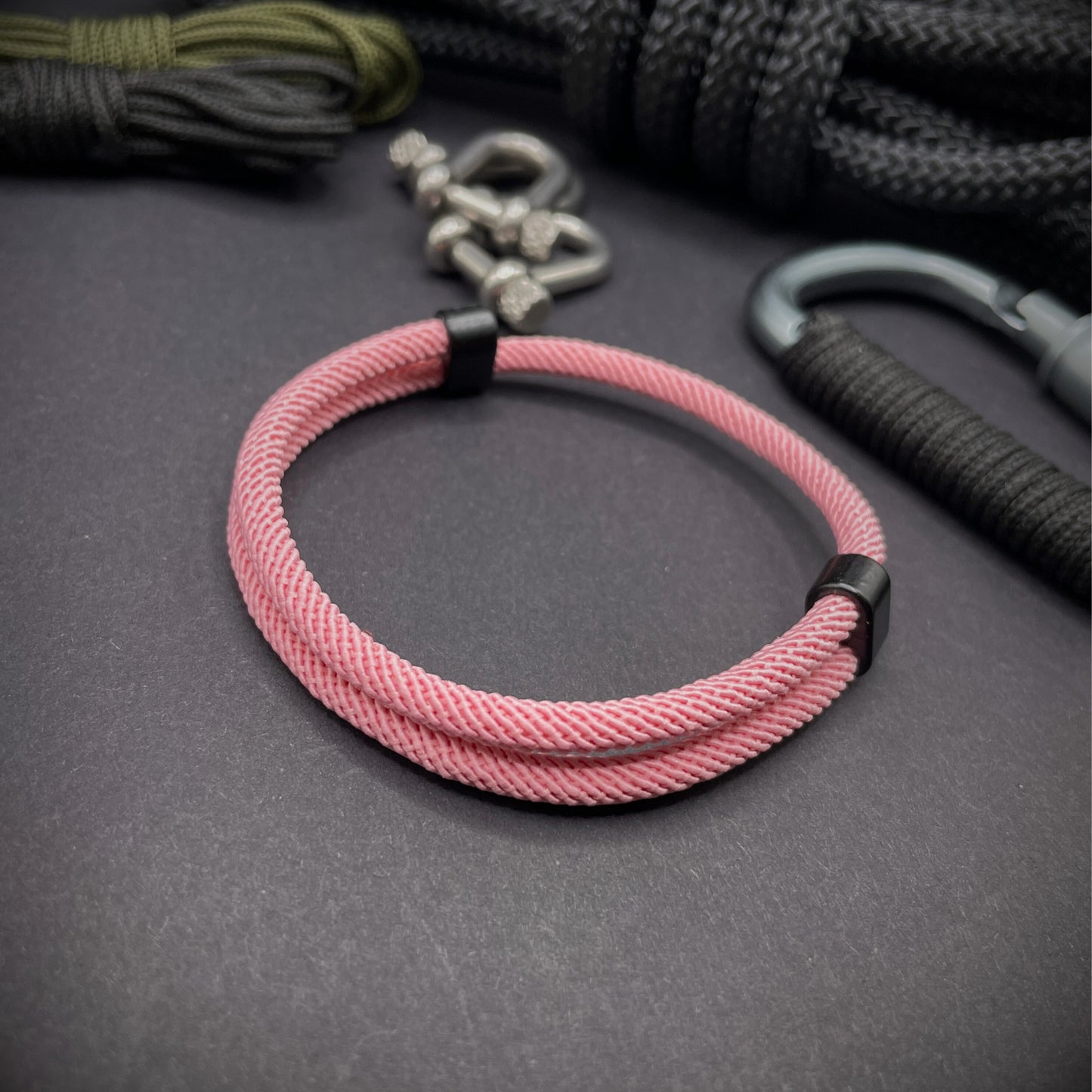 Pink Milan rope bracelet with black sliding clasps, displayed on a dark grey surface with EDC gear in the background.