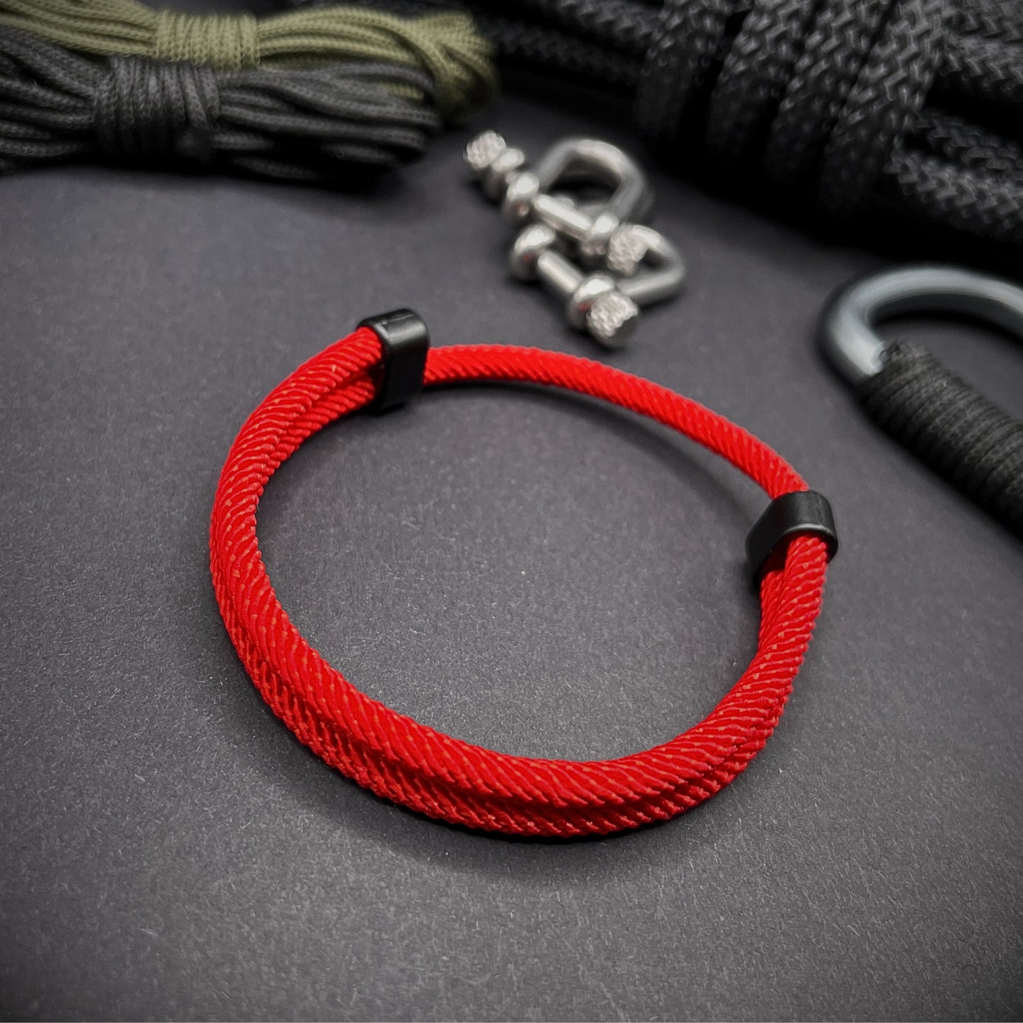 Red Milan rope bracelet with black sliding clasps, displayed on a dark grey surface with EDC gear in the background.