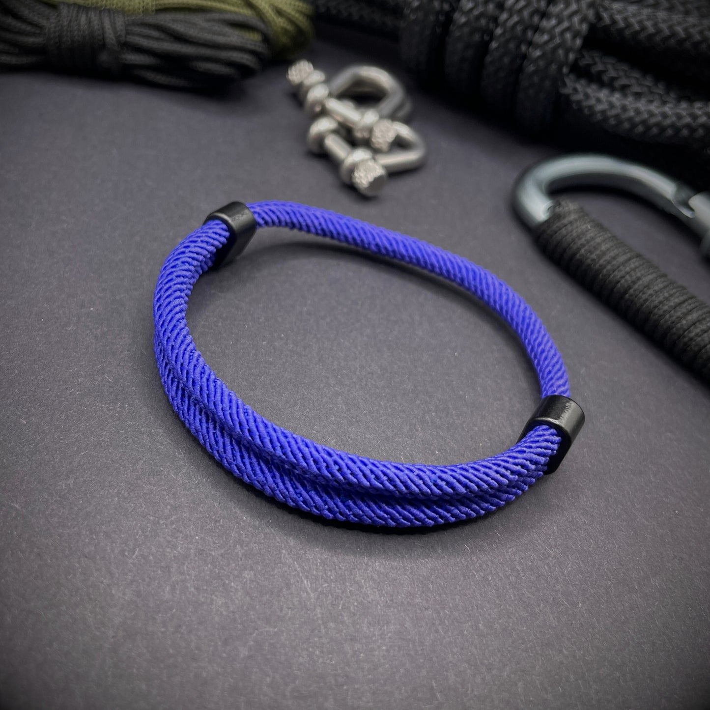 Royal Blue Milan rope bracelet with black sliding clasps, displayed on a dark grey surface with EDC gear in the background.