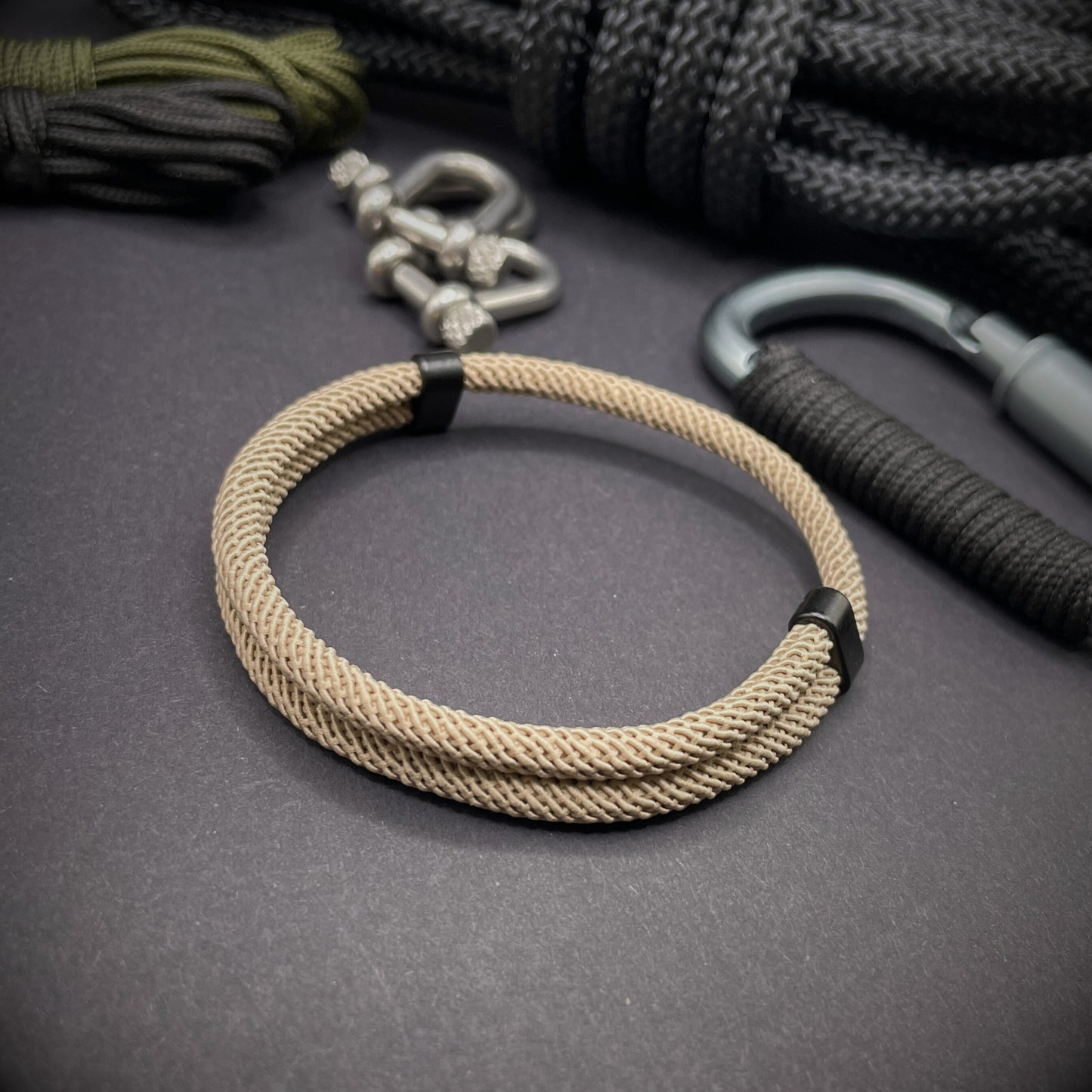 Tan Milan rope bracelet with black sliding clasps, displayed on a dark grey surface with EDC gear in the background.