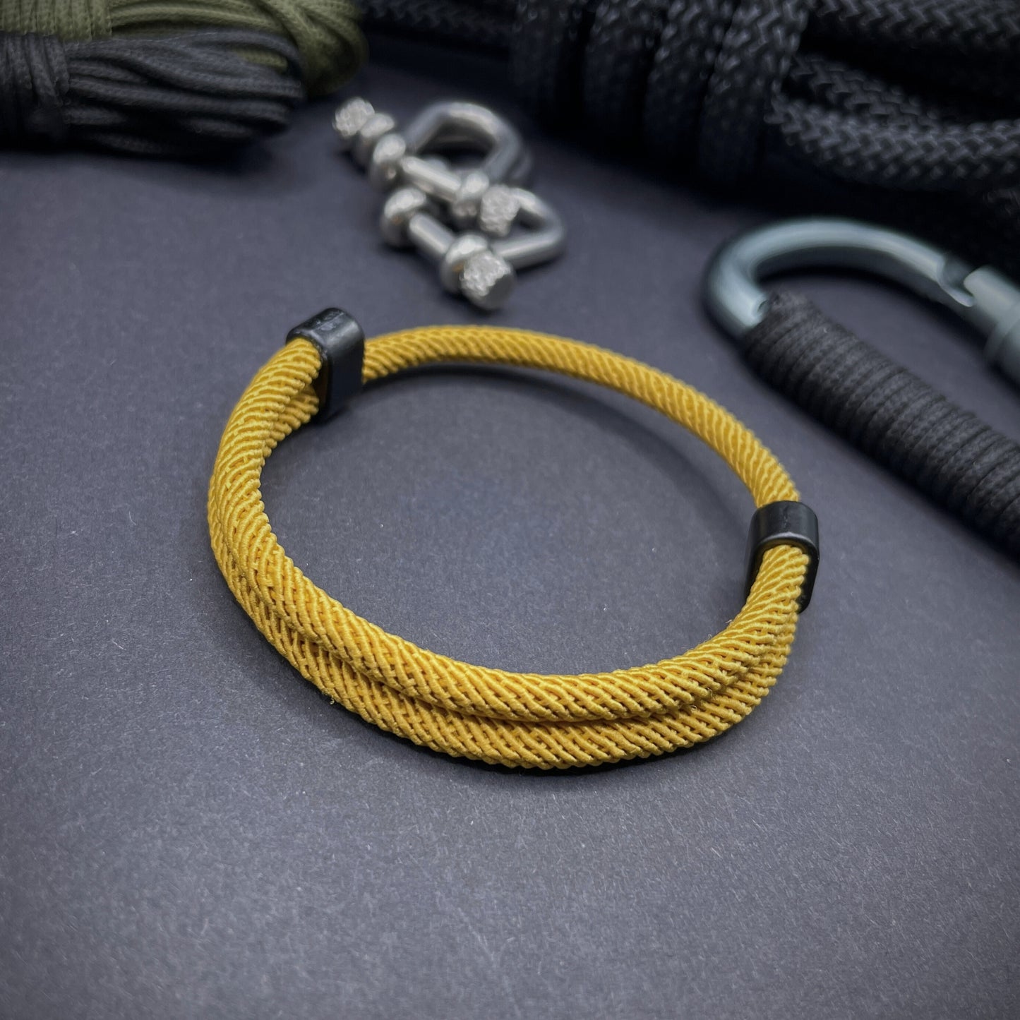 Turmeric Milan rope bracelet with black sliding clasps, displayed on a dark grey surface with EDC gear in the background.