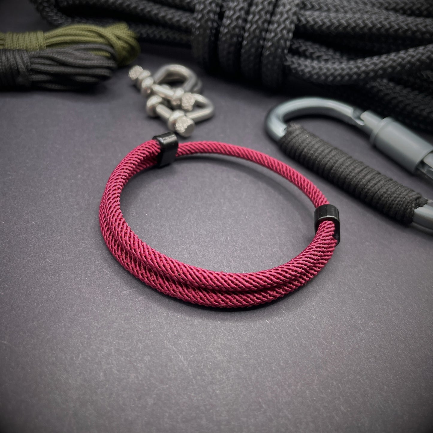 Wine Red Milan rope bracelet with black sliding clasps, displayed on a dark grey surface with EDC gear in the background.