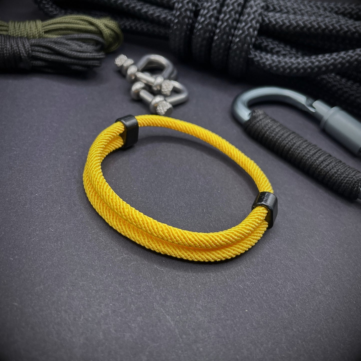 Yellow Milan rope bracelet with black sliding clasps, displayed on a dark grey surface with EDC gear in the background.