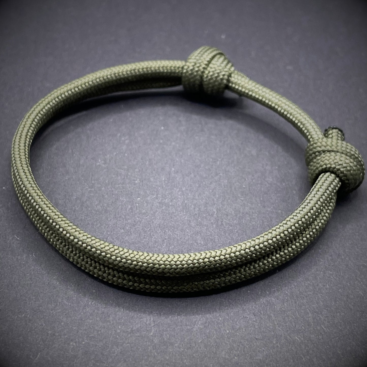 An army green paracord rope bracelet arranged on a dark grey surface.