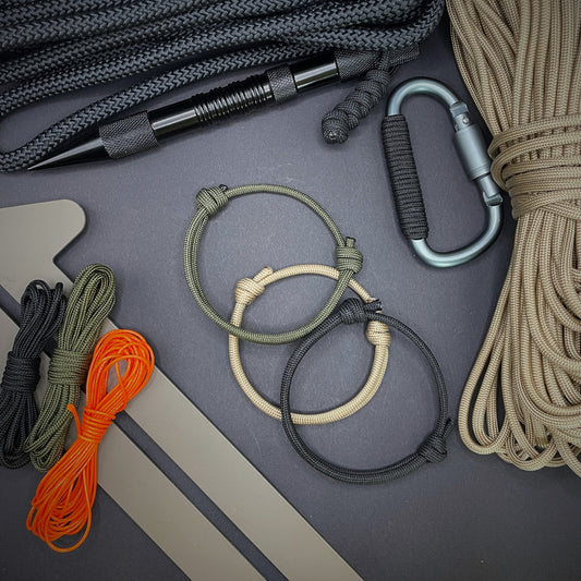 A collection of three paracord rope bracelets arranged on a dark grey surface and surrounded by EDC tools and spools of paracord.
