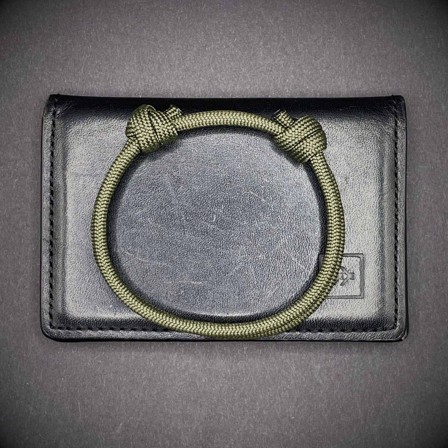 An army green paracord rope bracelet arranged on a dark grey surface.