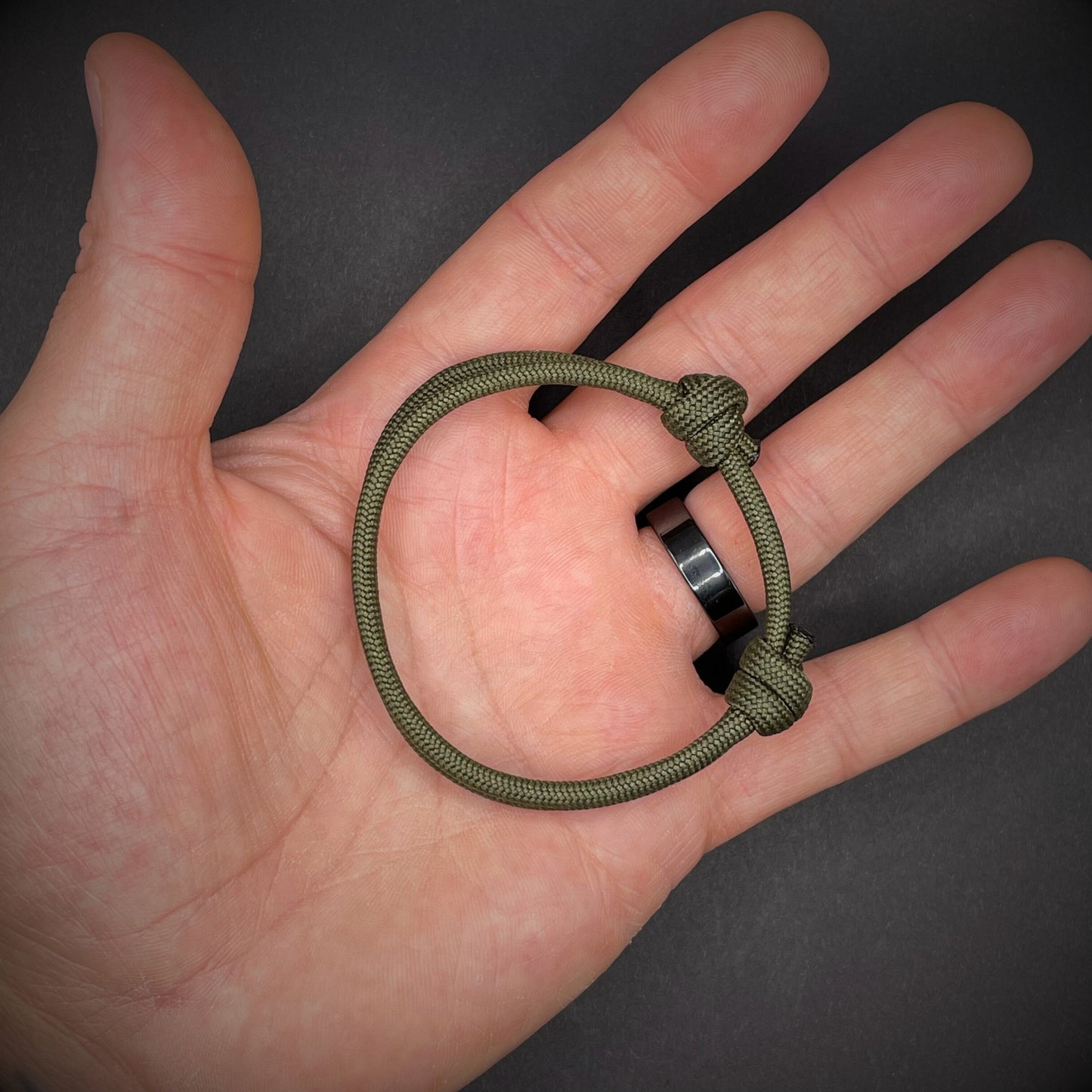 An army green paracord rope bracelet arranged in an open palmed hand over a dark grey surface.