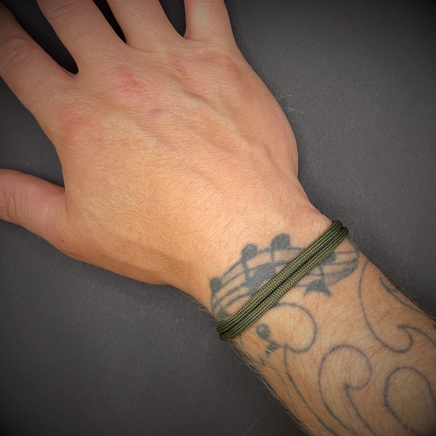 An army green paracord rope bracelet on a tattooed wrist over a dark grey surface.