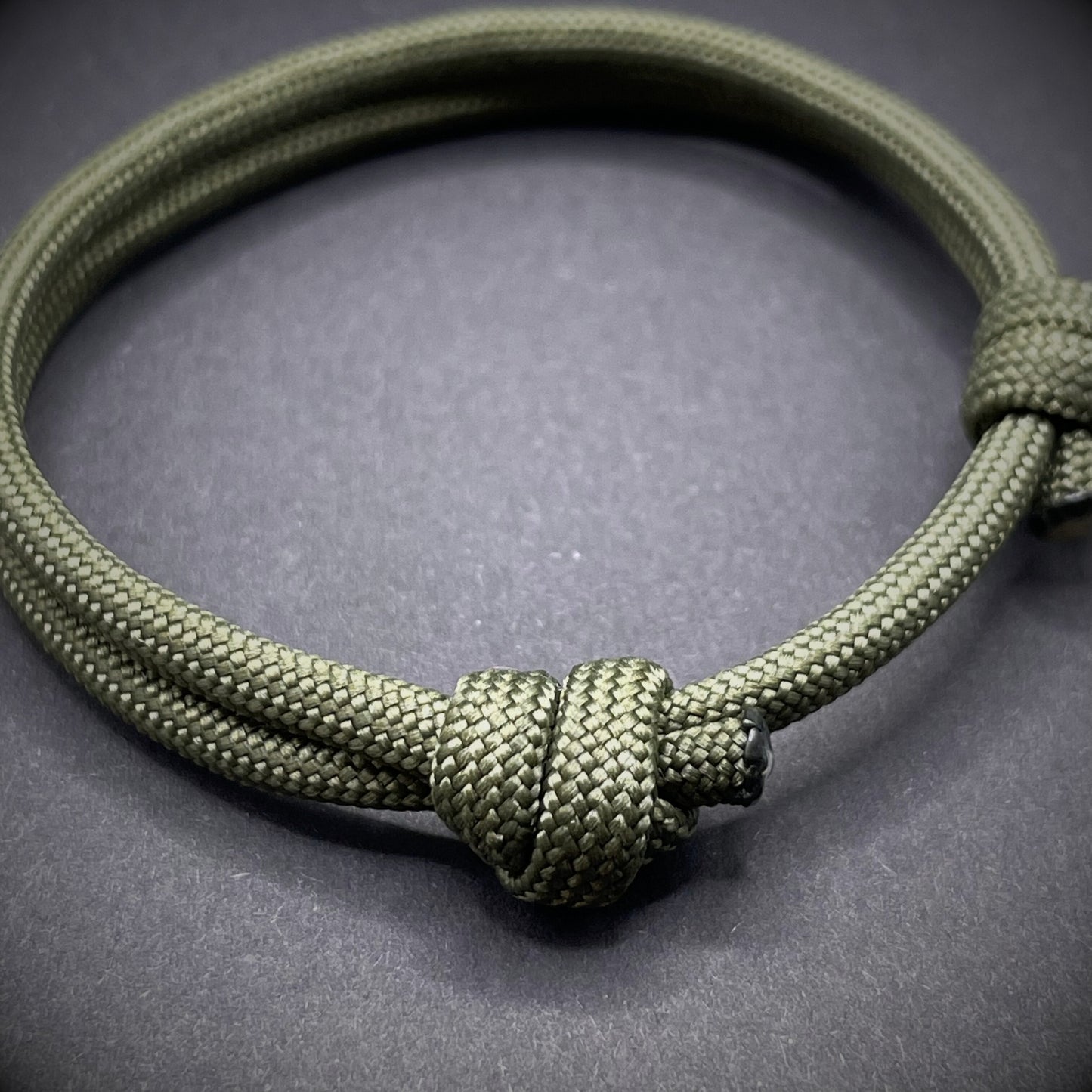 An army green paracord rope bracelet arranged on a dark grey surface.
