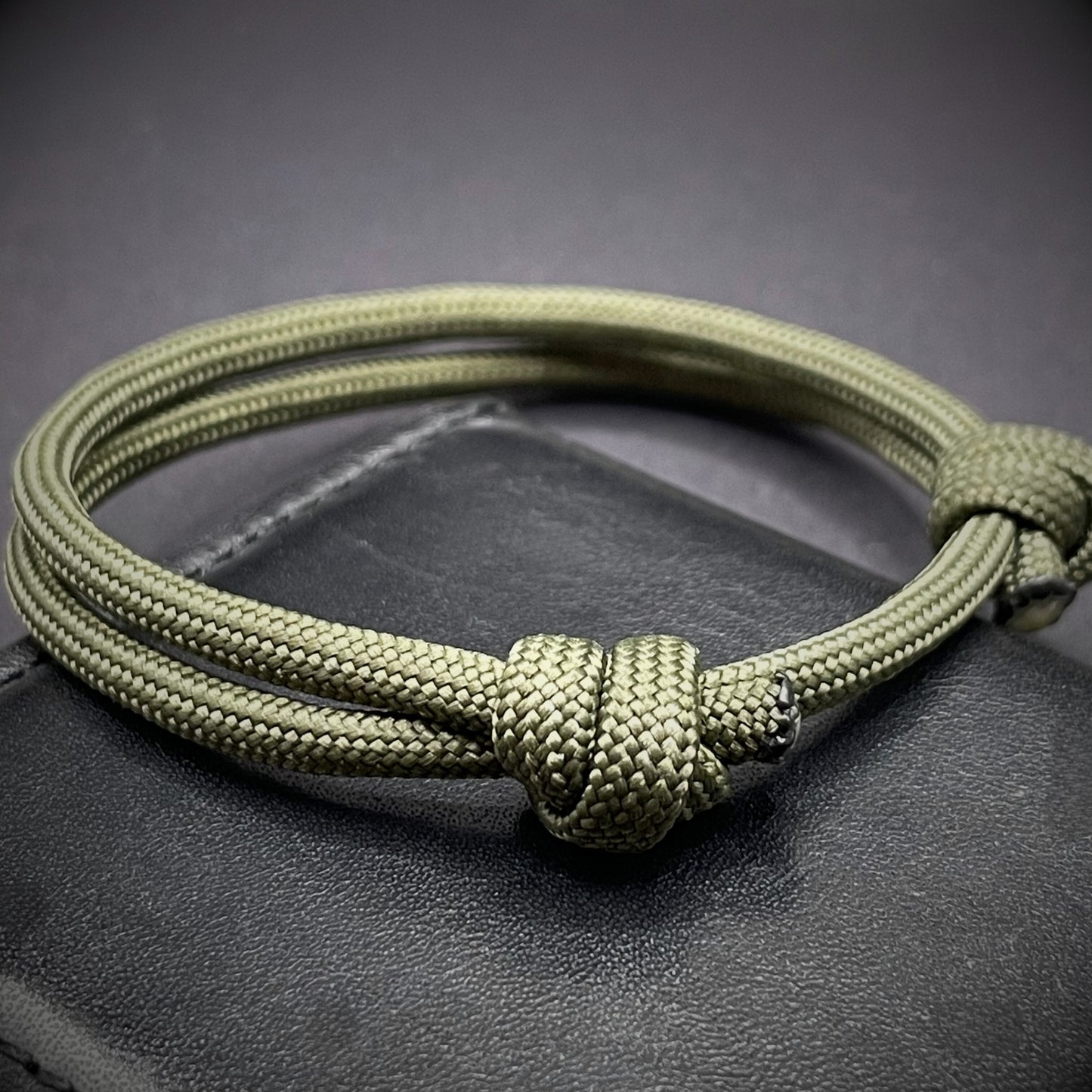 An army green paracord rope bracelet arranged on a dark grey surface.