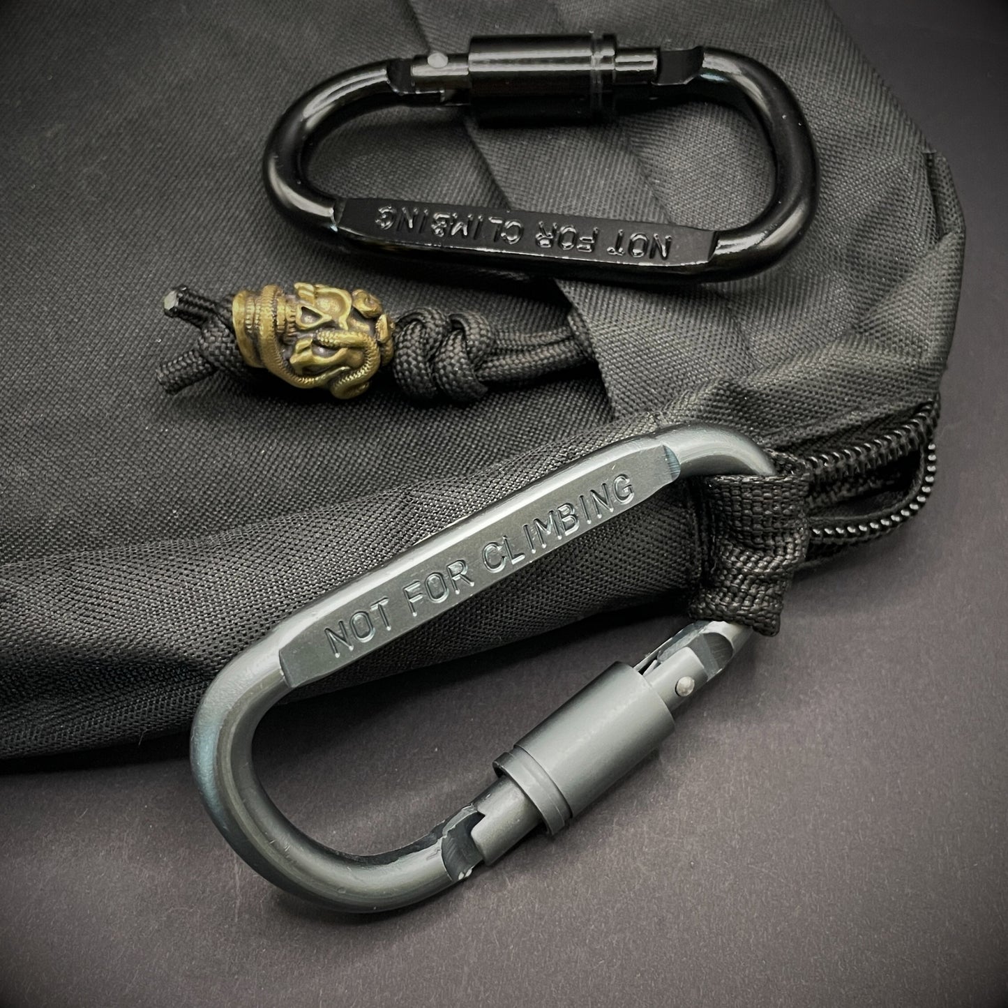 Two carabiners marked 'NOT FOR CLIMBING' clipped to a black pouch, one with a brass bead on paracord.