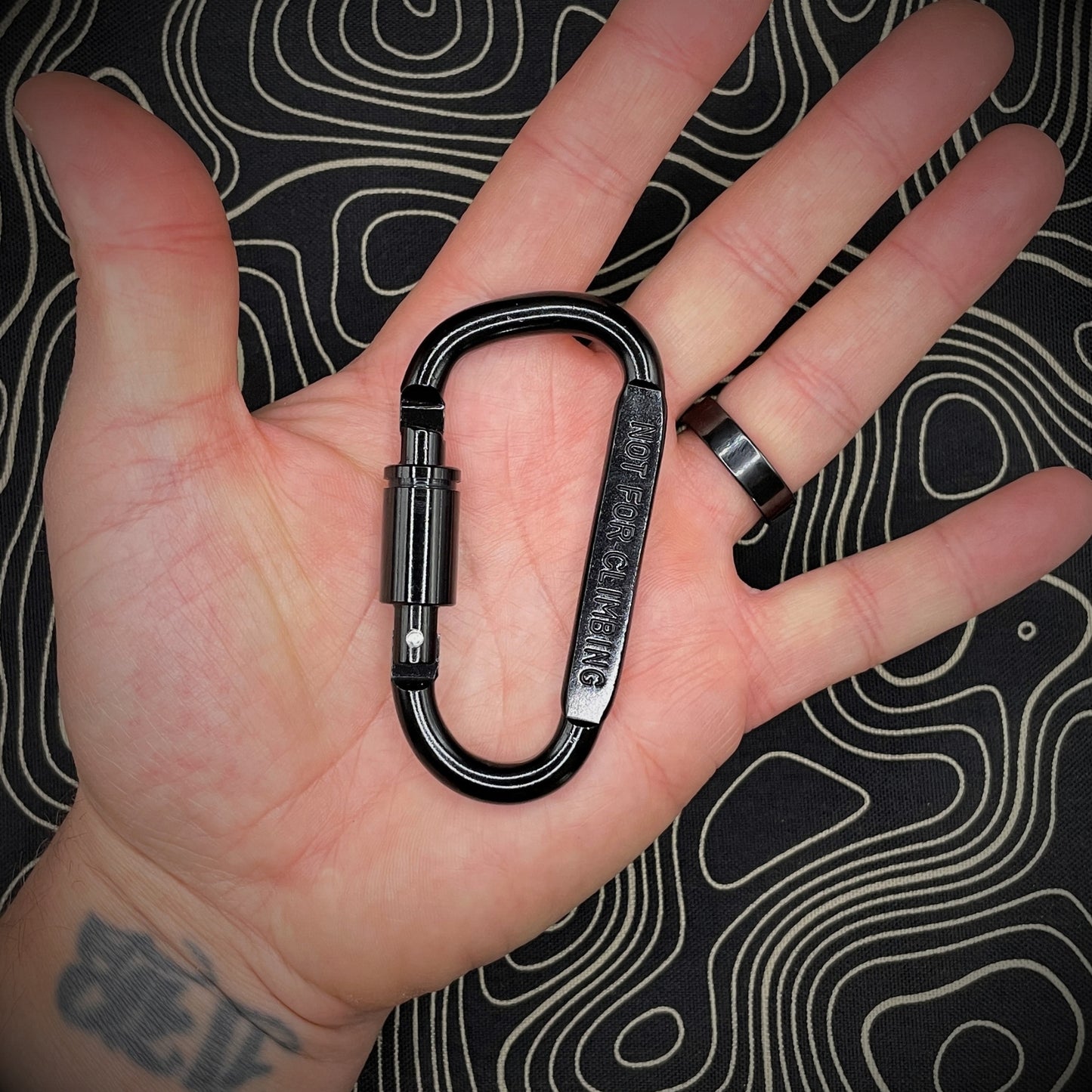 Hand holding a black carabiner marked 'NOT FOR CLIMBING' against a black background with topographic line pattern.