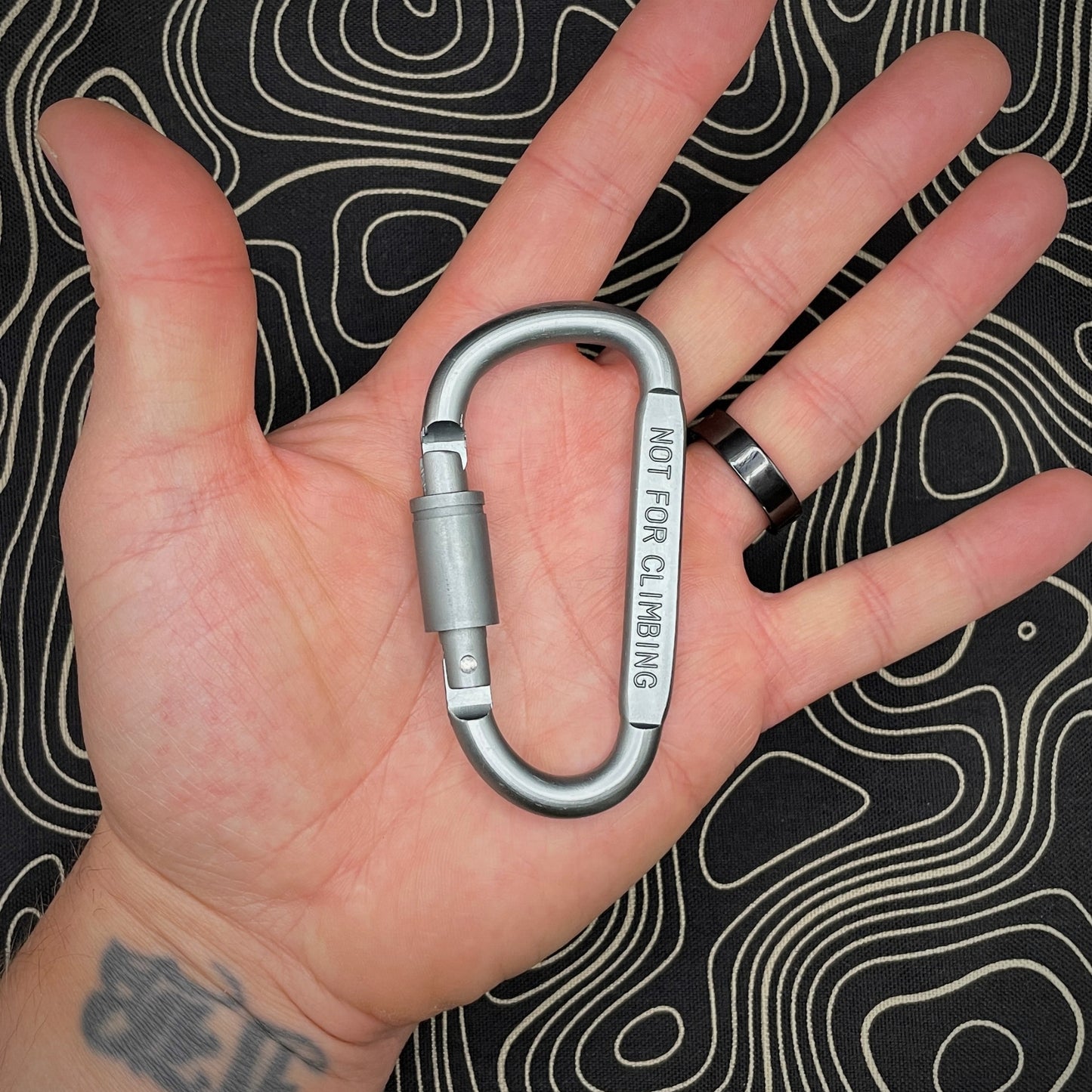 Hand holding a grey carabiner marked 'NOT FOR CLIMBING' against a black background with topographic line pattern.