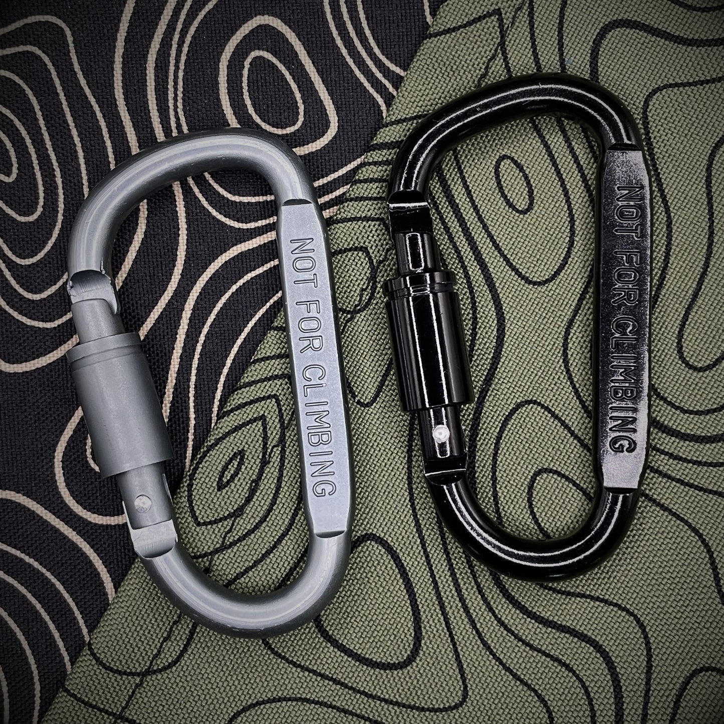 Black and grey carabiners marked 'NOT FOR CLIMBING' against a black background with topographic line pattern.