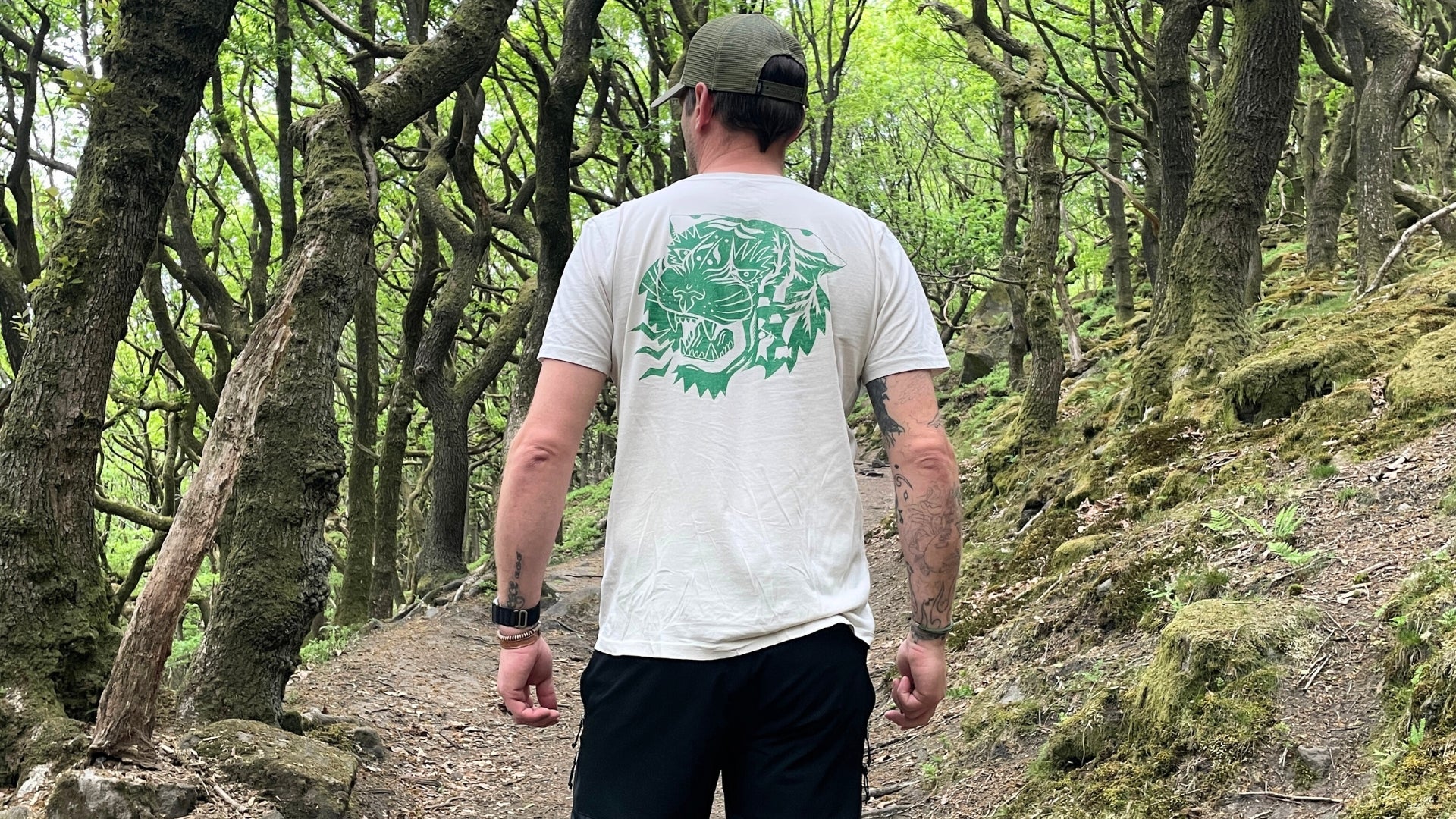 An man wears an Oat coloured t-shirt with a green tiger design printed on the back whilst walking in woodland.