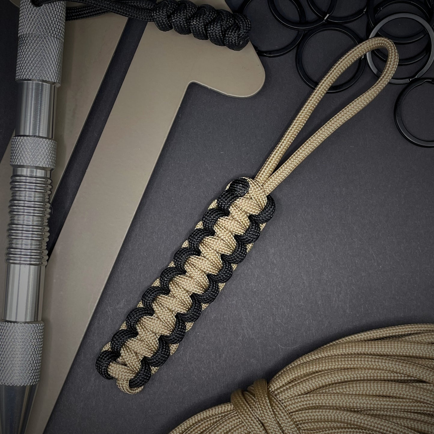 Flat lay of Khaki and Black 550 Paracord knife lanyard on a dark grey surface surrounded by EDC items