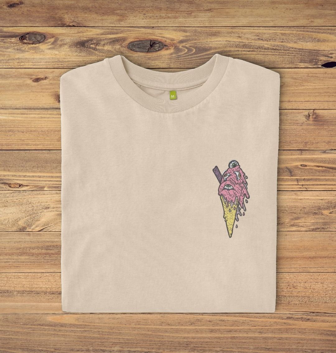 Folded Oat coloured t-shirt on a oak panelled background, the t-shirt has an ice cream with eyes in as a logo print.