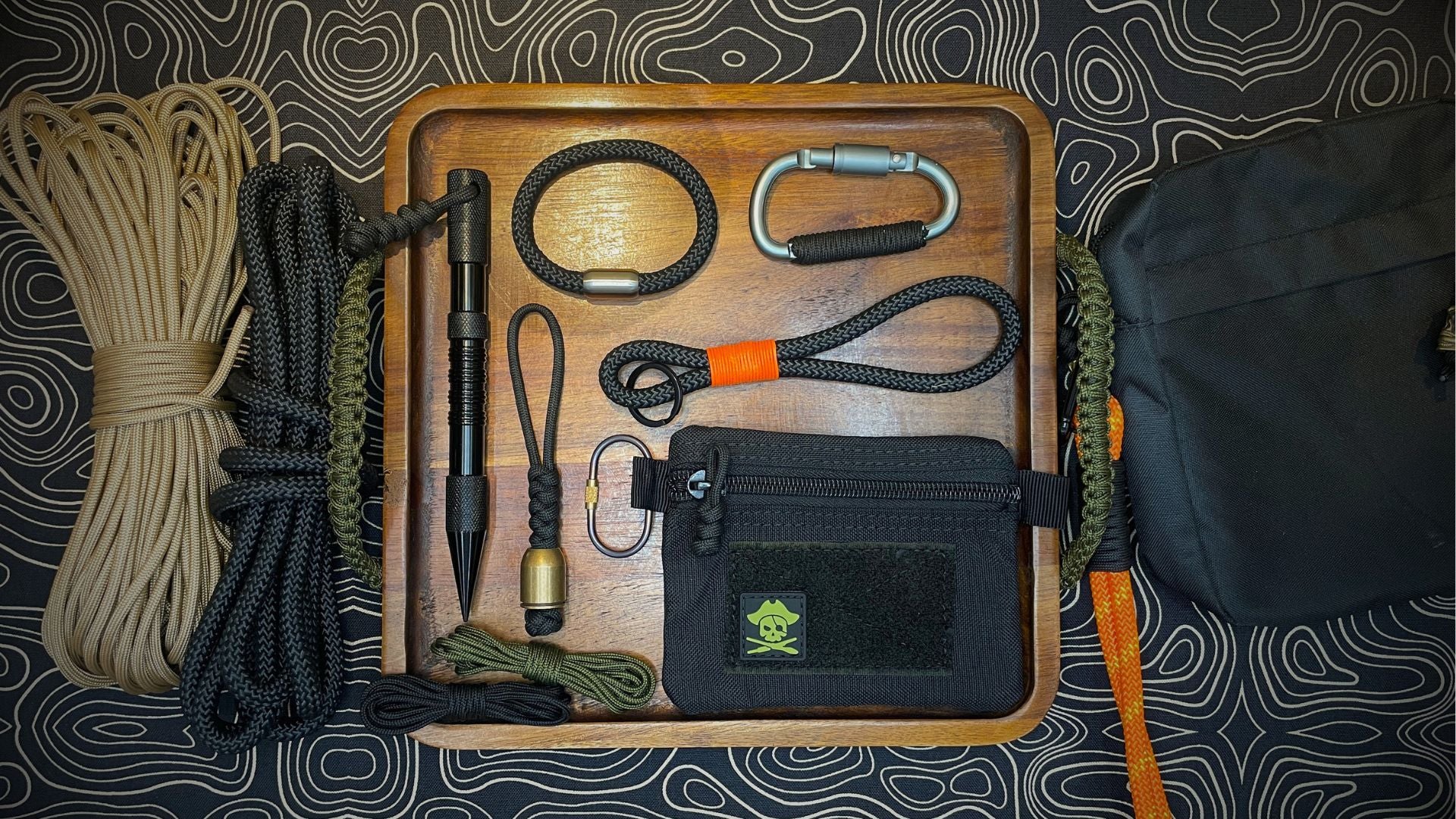 Tray with paracord accessories, lanyards, carabiner, EDC pen, and pouch, all laid out on a topographic-patterned cloth.