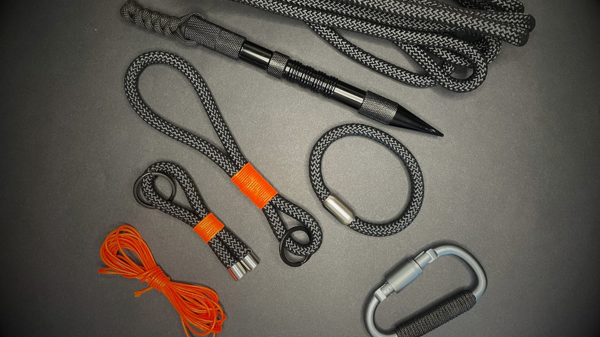 Flat lay of black and grey EDC accessories including a tactical pen, paracord loops with orange whipping, bracelet, carabiner, and orange utility cord on a dark surface.