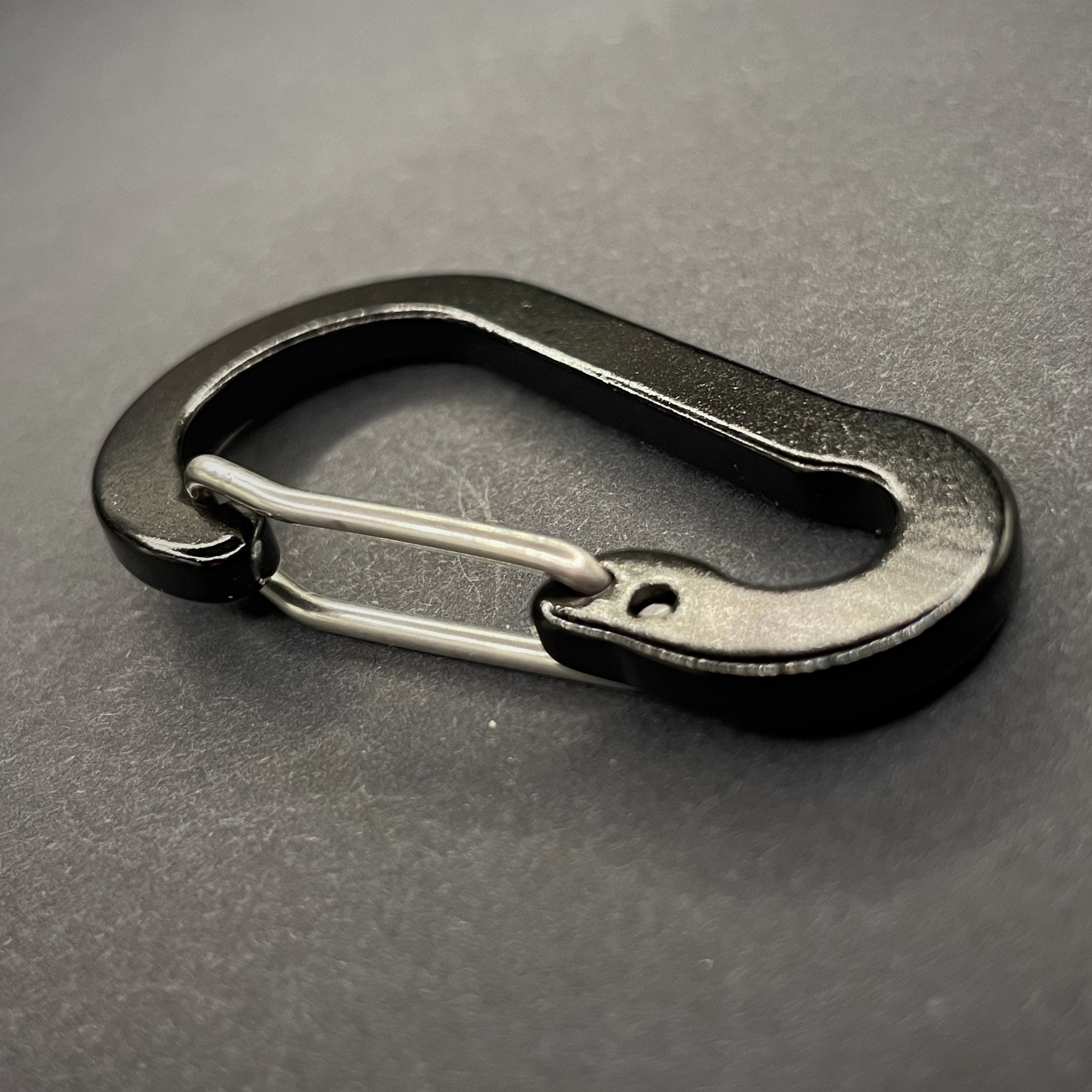 An angled view of a Black Anodised aluminium carabiner, arranged on a dark surface
