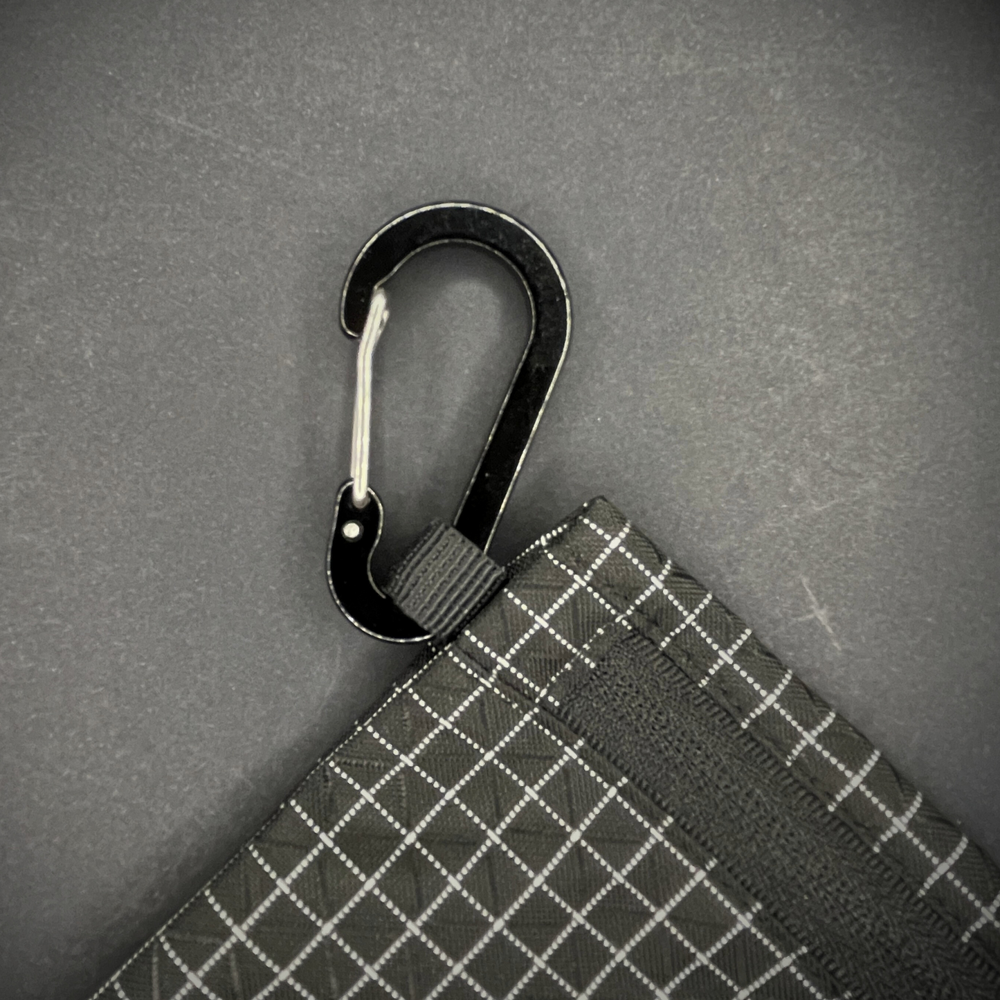 Black aluminium carabiner attached to a black grid-patterned pouch, arranged on a dark surface.
