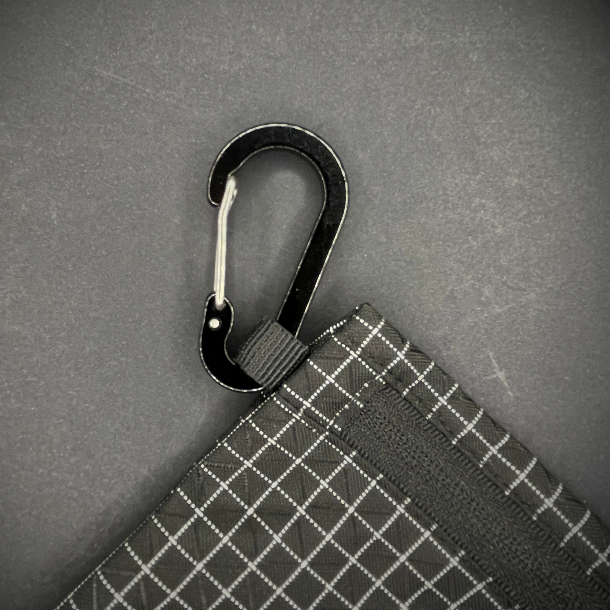 Black aluminium carabiner attached to a black grid-patterned pouch, arranged on a dark surface.
