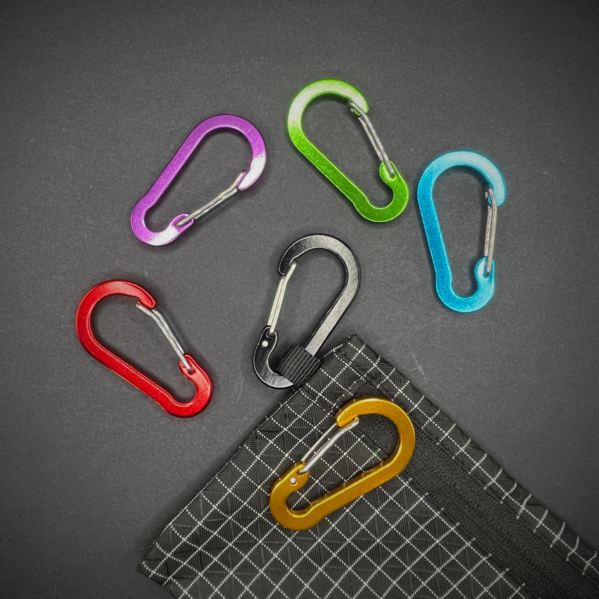 Six colourful aluminium carabiners and one attached to a black grid-patterned pouch, arranged on a dark surface.